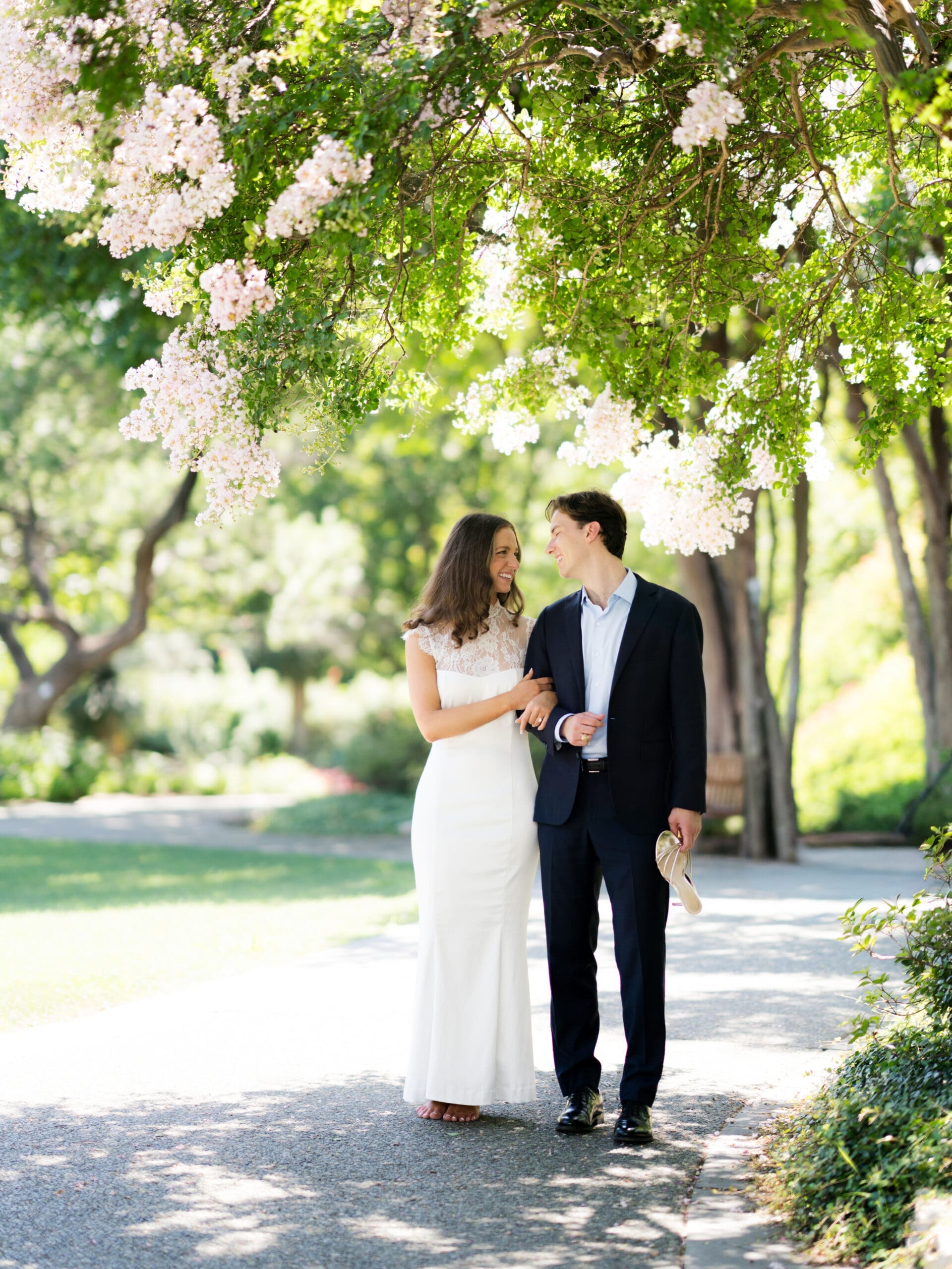 Couple walking hand in hand beneath blossoming pink flowers on a garden path.