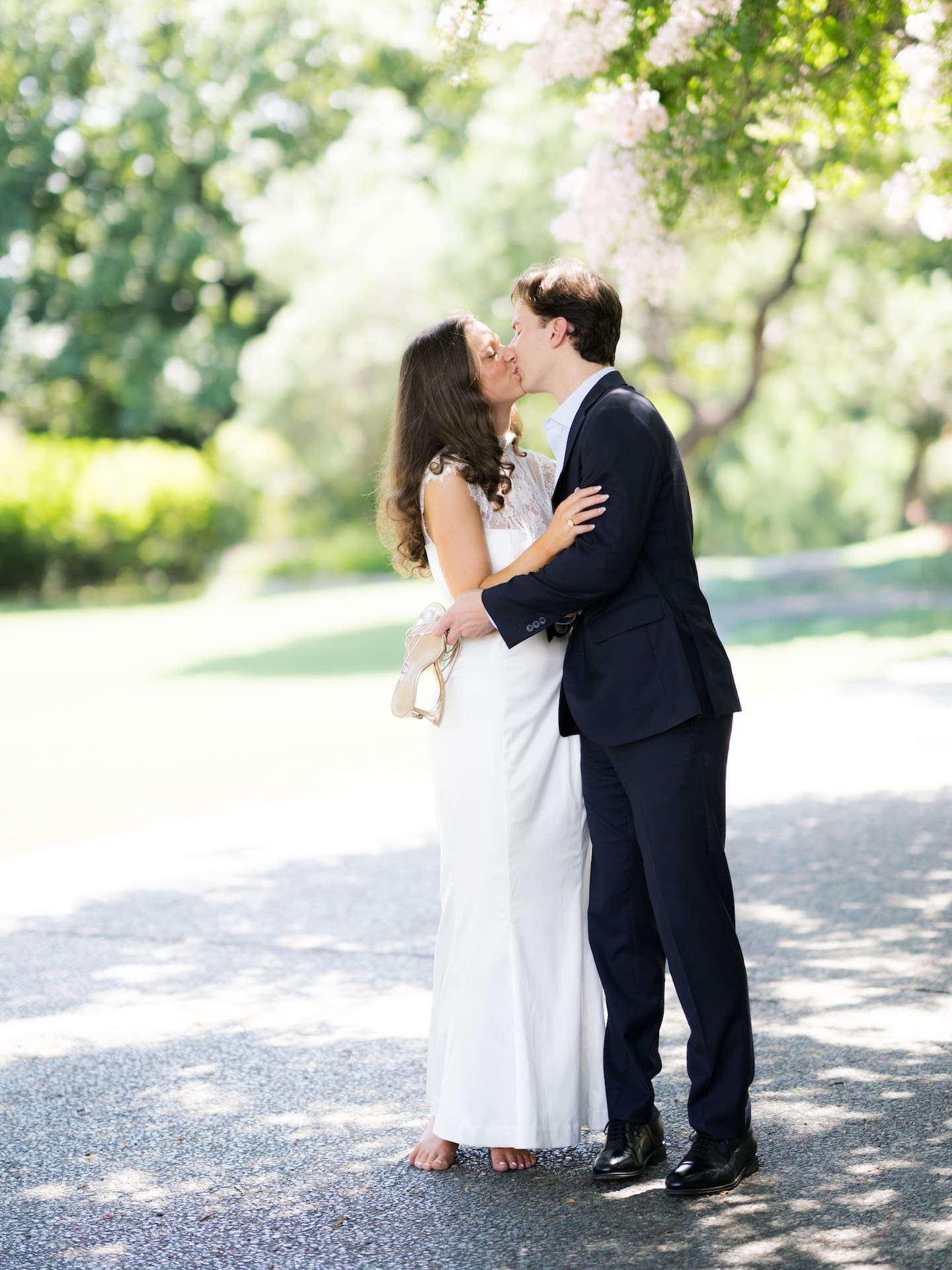 Fall engagement photos of a couple kissing on a sunlit path shaded by blossoming trees.