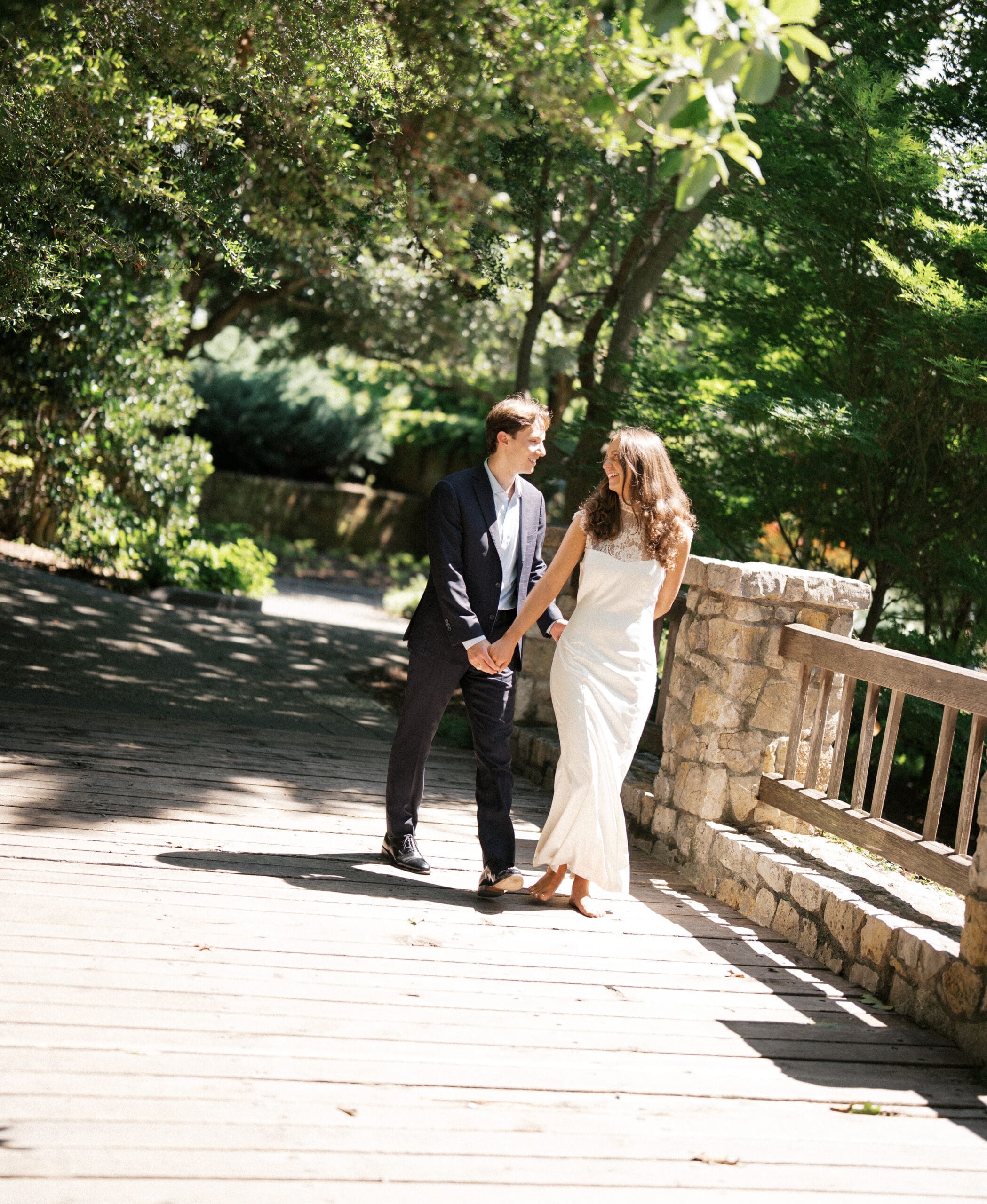 Couple walking hand in hand and smiling on a wooden garden bridge.