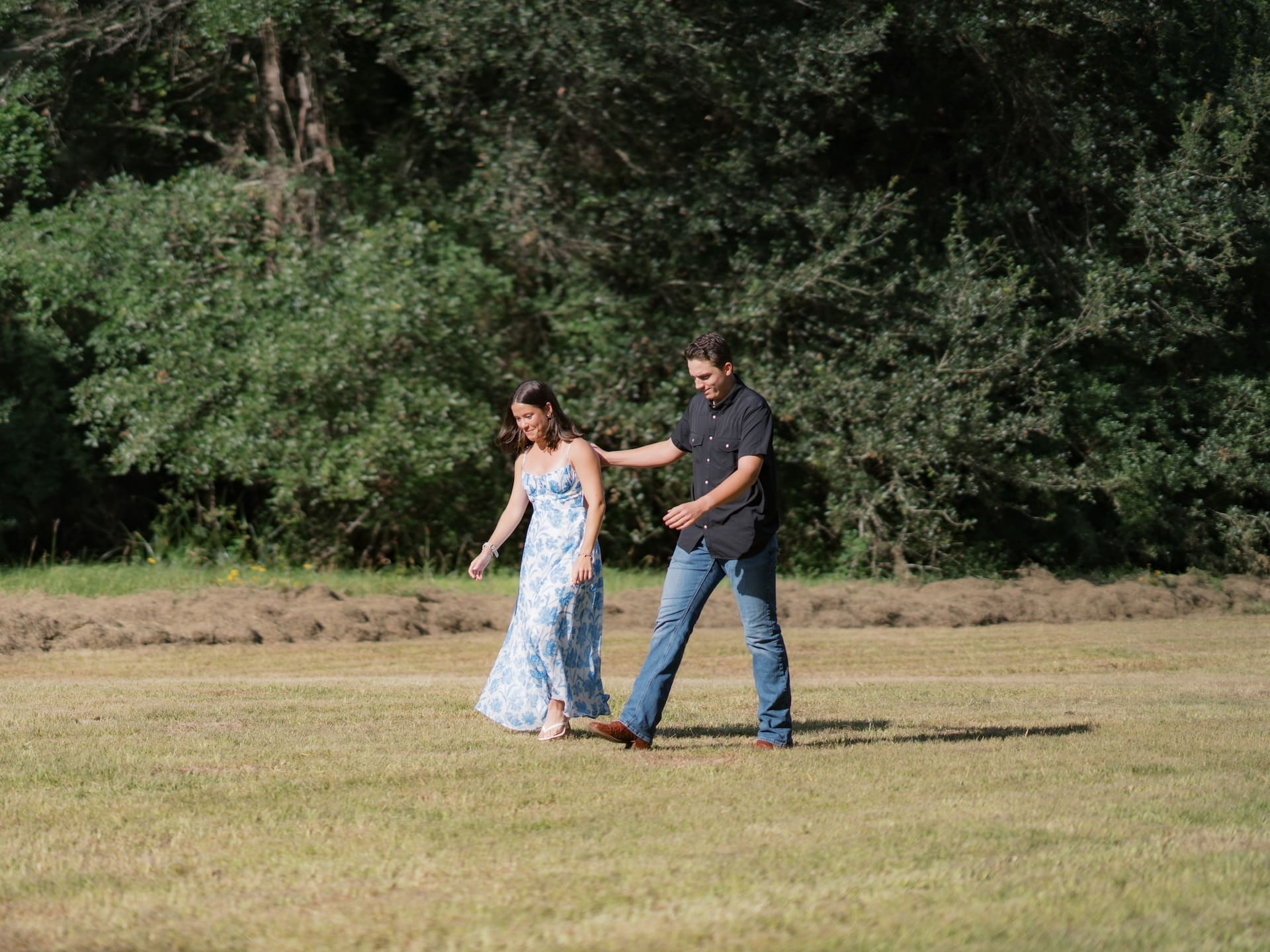 Man guiding his girlfriend by the shoulder as they walk together across a sunny field.