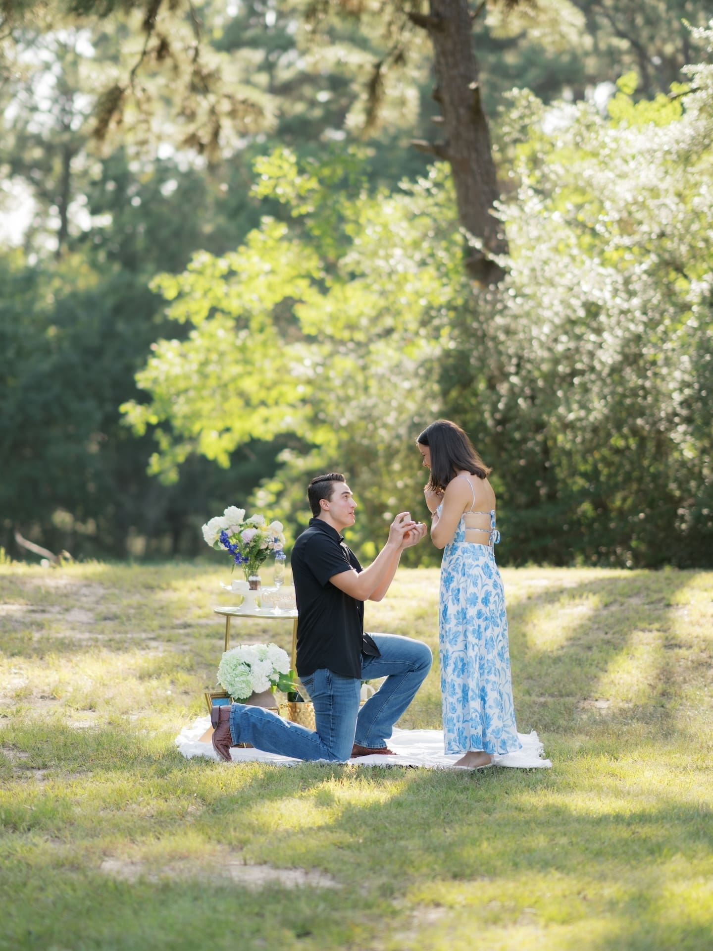 Man kneeling to propose to his girlfriend under blossoming trees in a sunlit field.