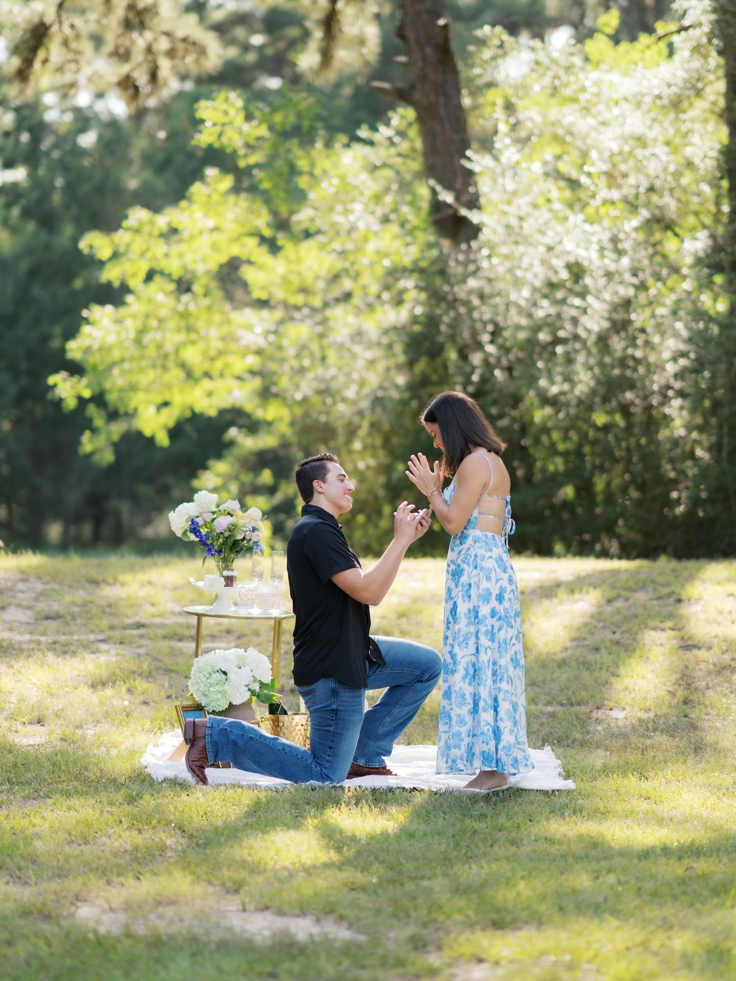 Man kneeling to propose to his girlfriend under blossoming trees in a sunlit field.