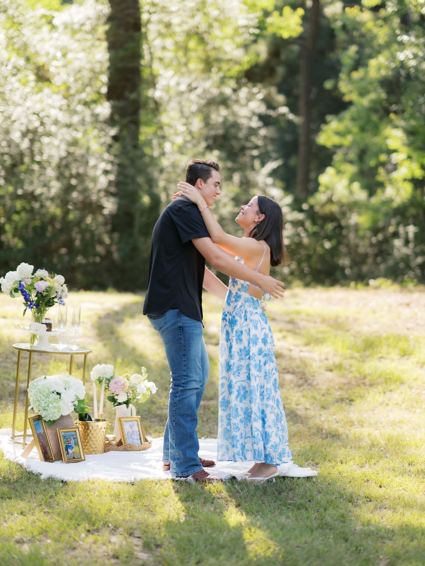 Newly engaged couple embracing joyfully beside a decorated picnic setup.