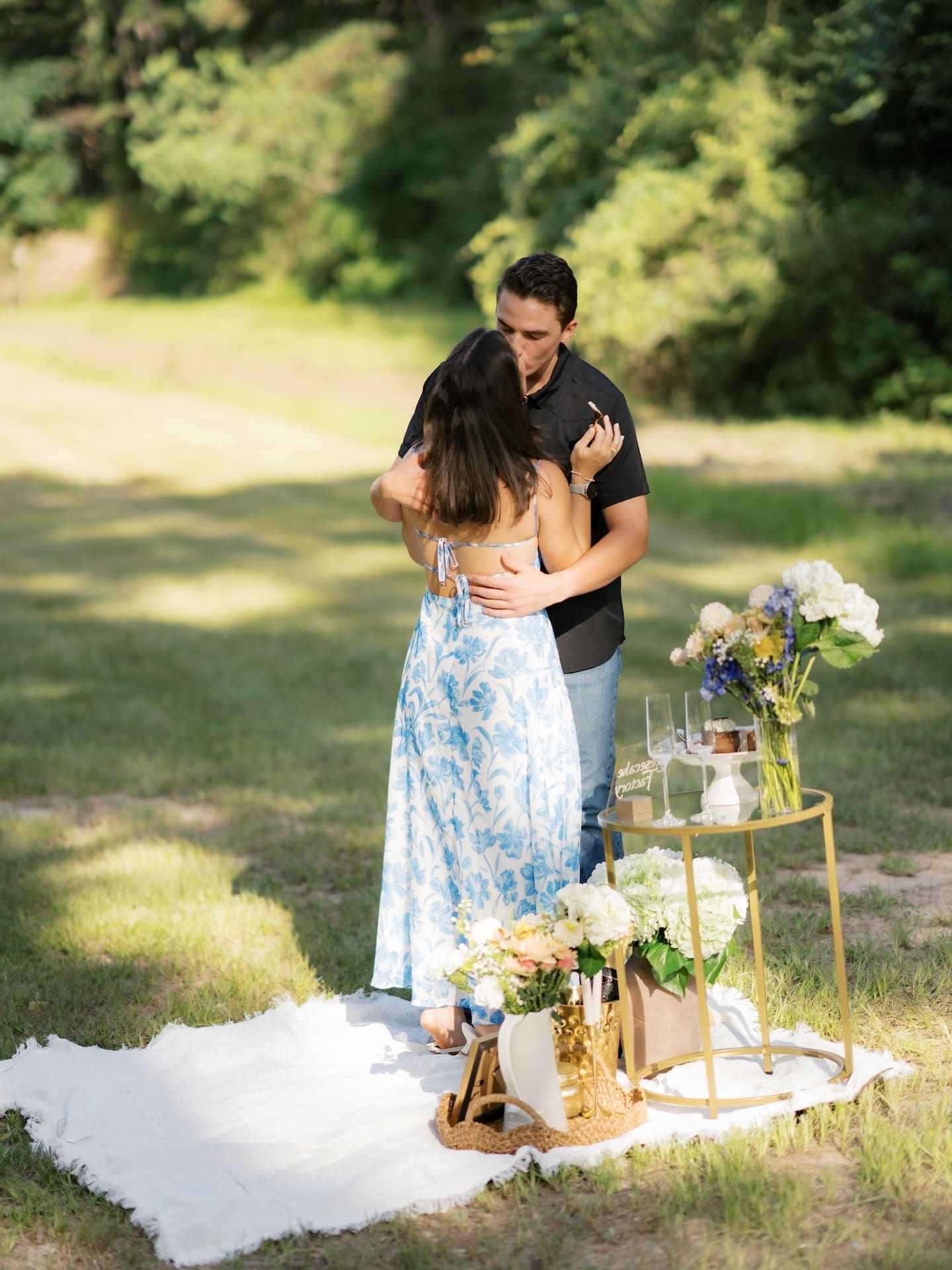 Newly engaged couple sharing a kiss beside a decorated picnic setup.