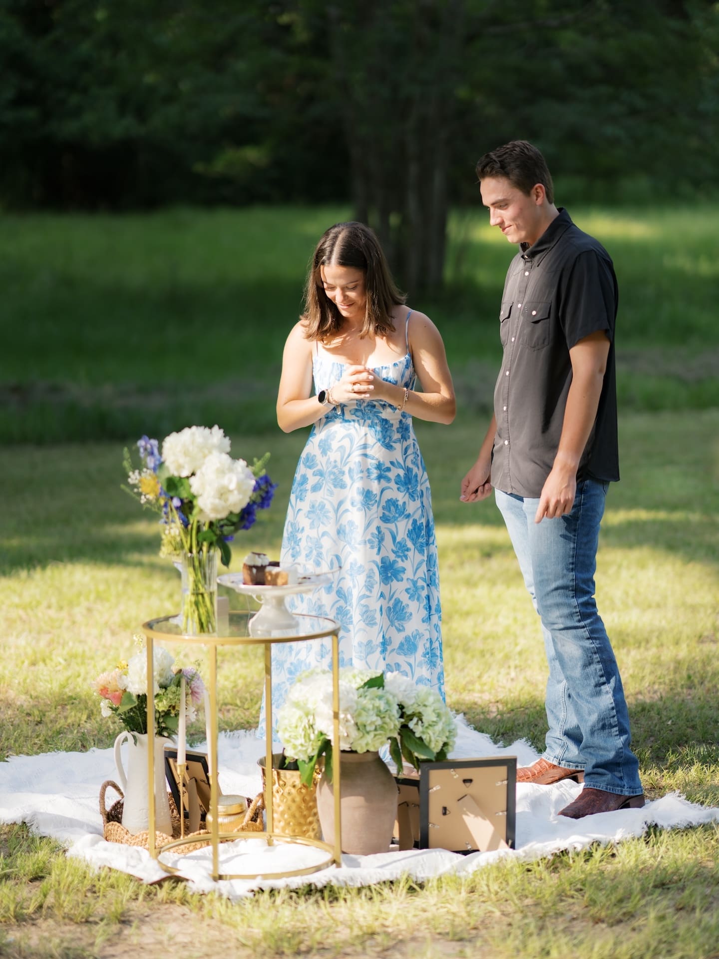 Couple looking down at a new engagement ring beside a floral picnic display.