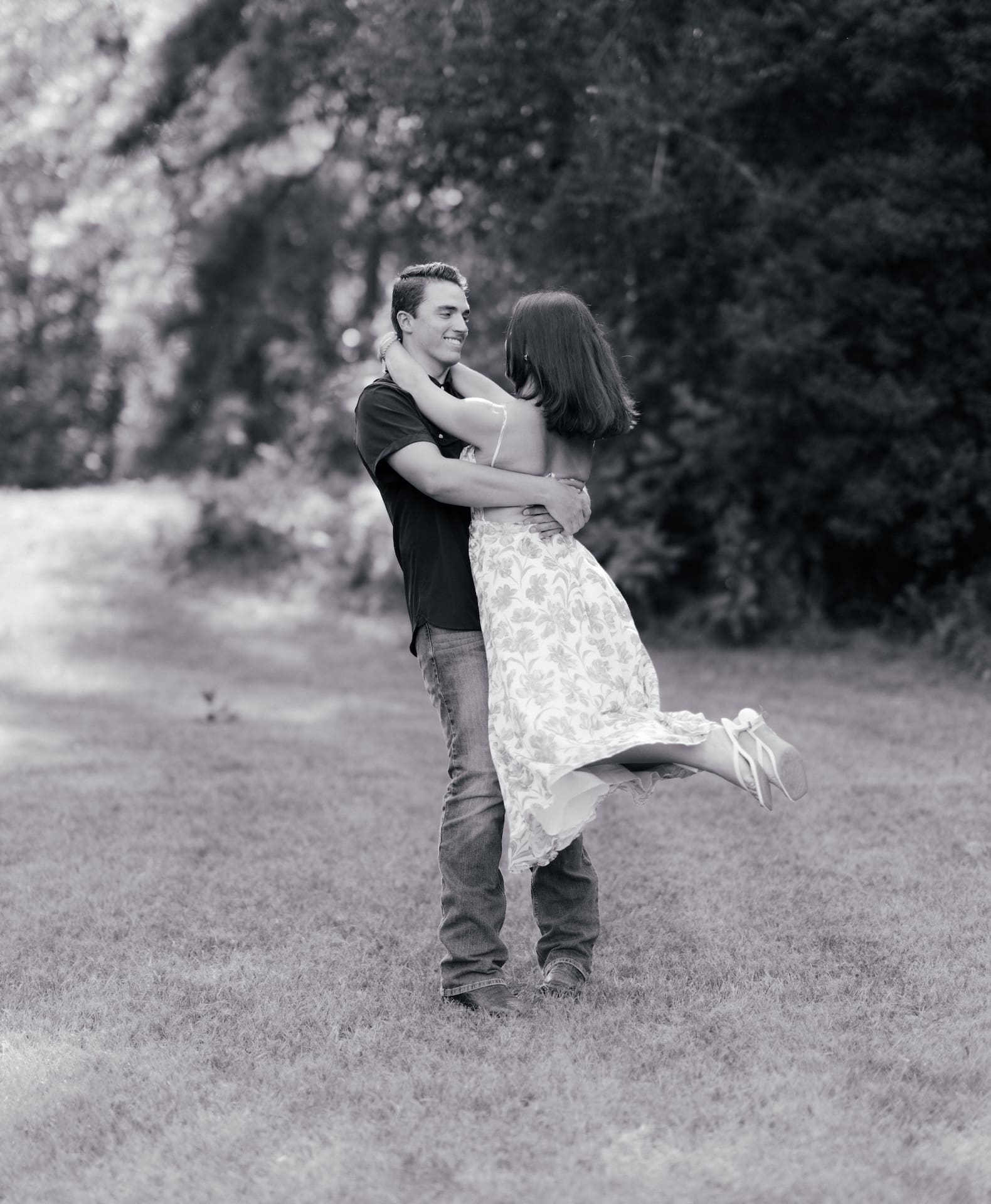 Black and white photo of a man lifting his fiancée playfully in a grassy field.