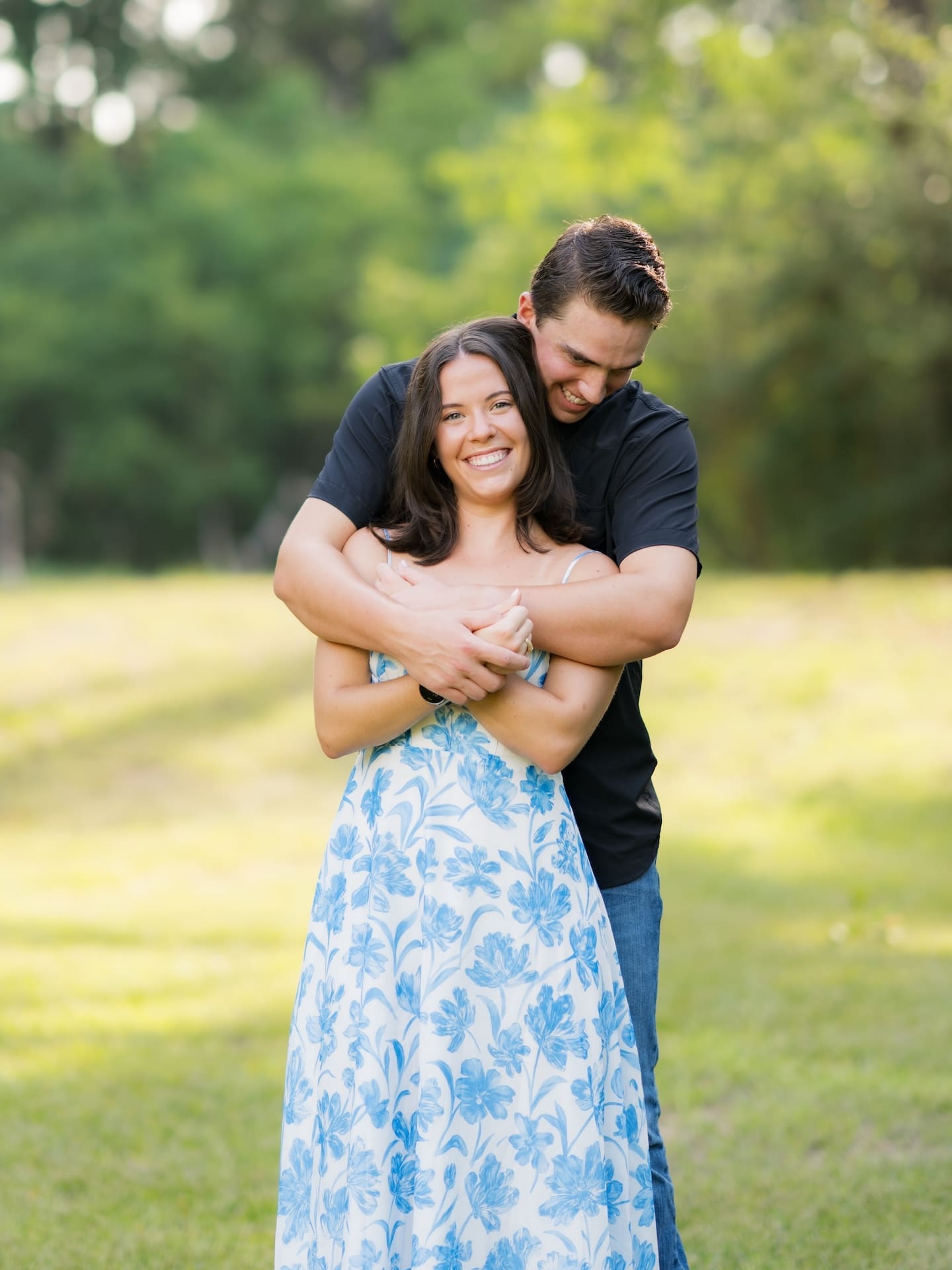 Man embracing his fiancée tightly from behind in a sunny grassy field.