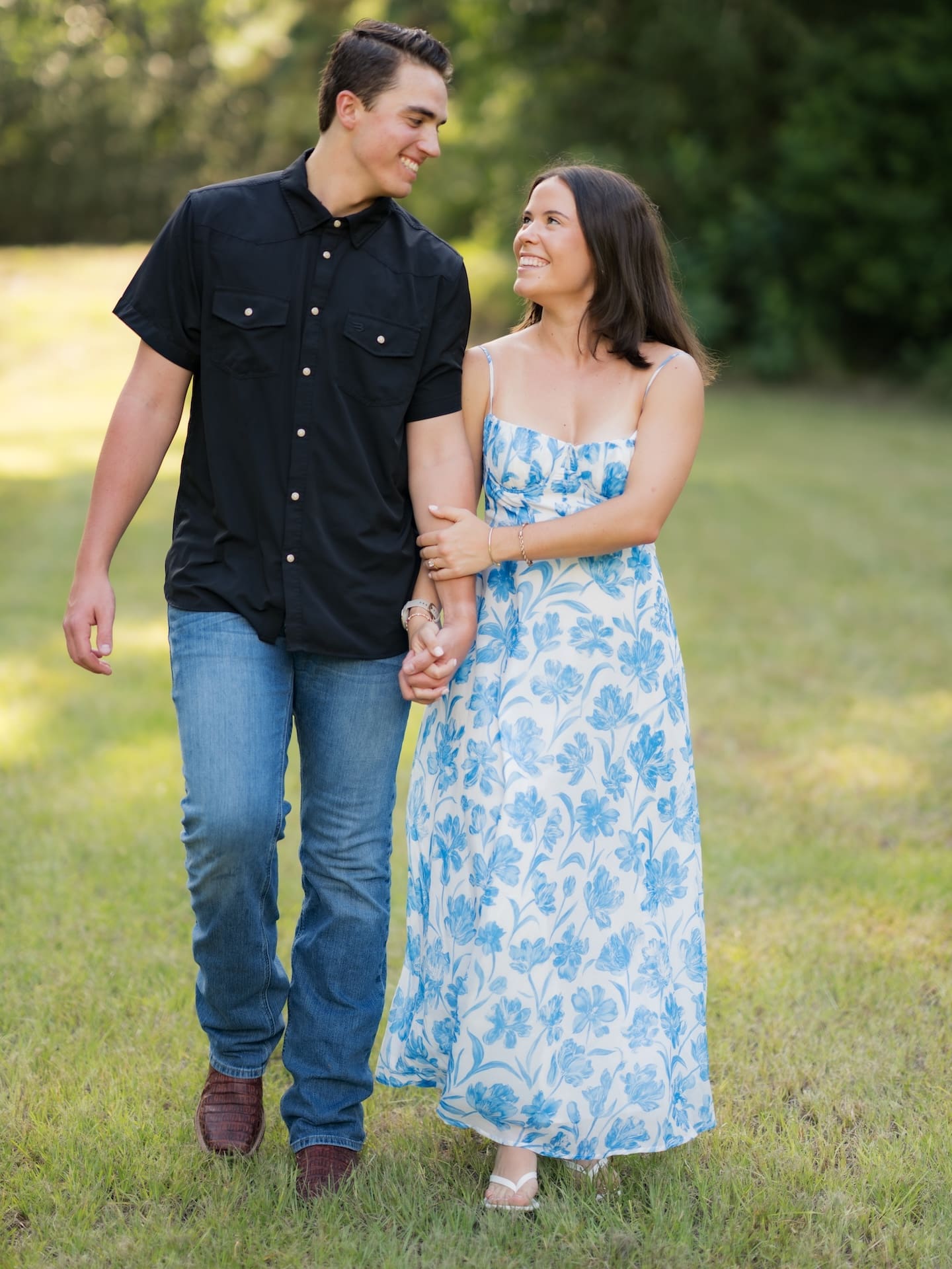 Newly engaged couple smiling at each other while holding hands on the grass.
