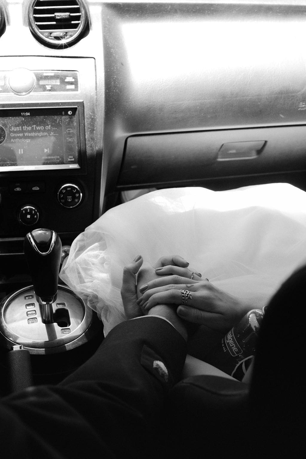 Black and white close-up inside the car, hands intertwined on her tulle skirt as "Just the Two of Us" plays on the dashboard screen.