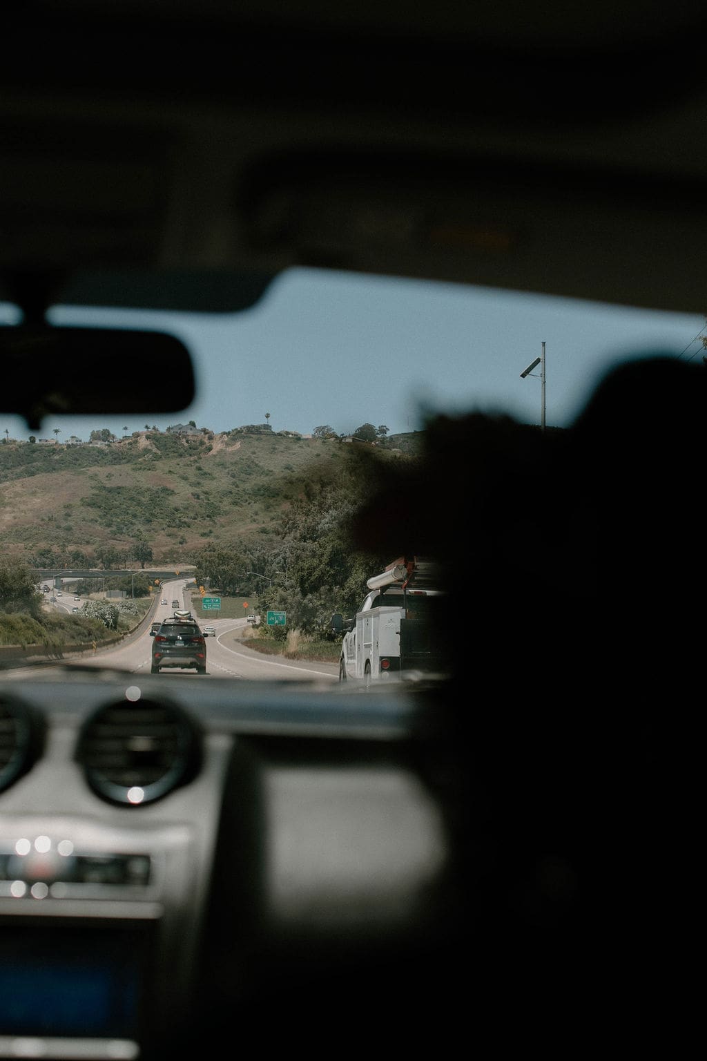 Candid shot from inside the moving car, a silhouette in the foreground as the highway and hills unfold through the windshield.