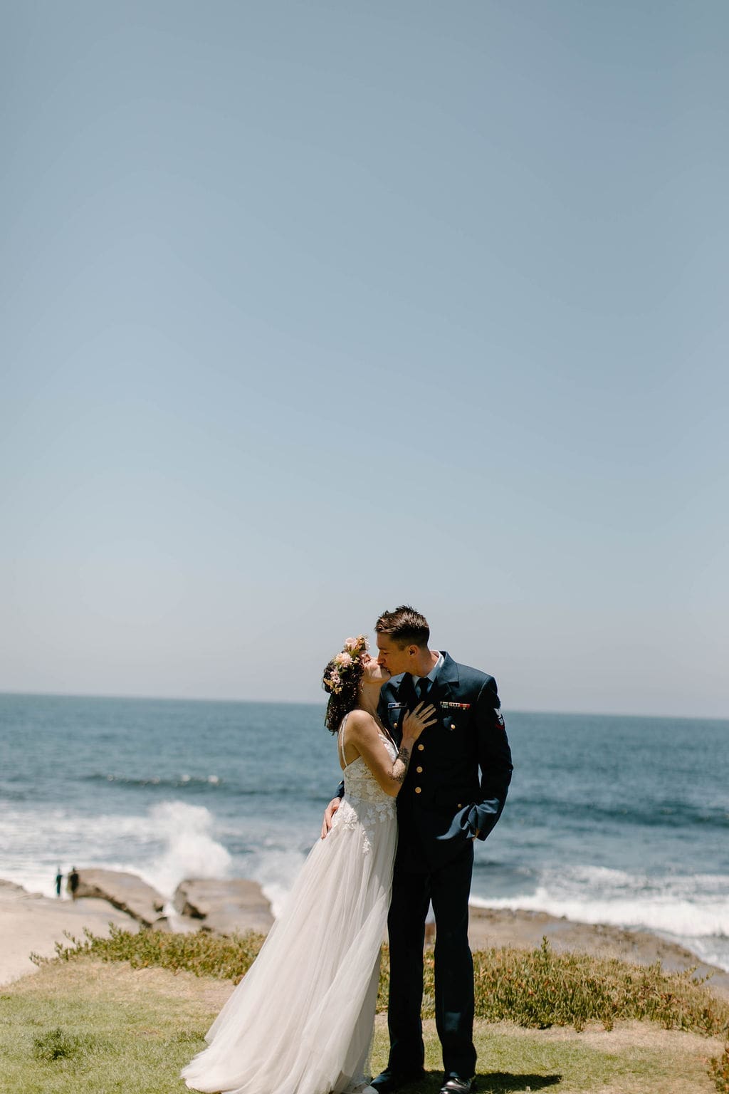 Bride laughs brightly with her head tipped back against her groom's shoulder, the ocean stretching out behind them.