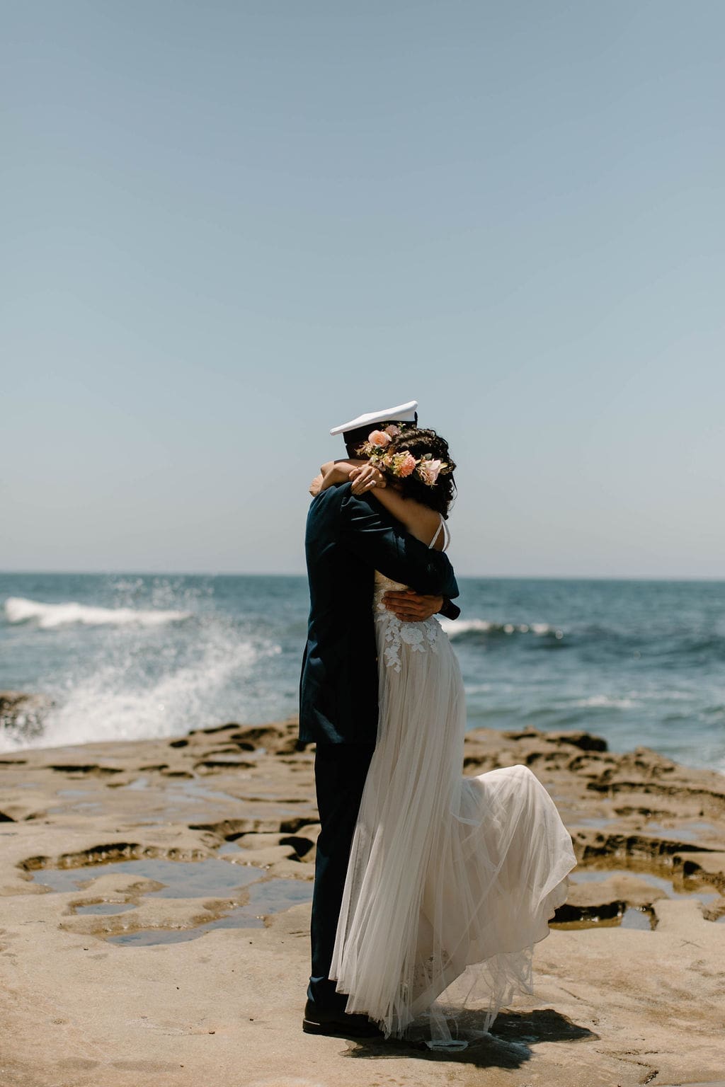 The couple wrapped in a tight embrace on the rocks, her flower crown pressed into his shoulder as waves crash beside them.