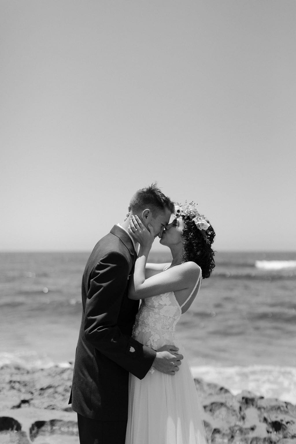 Black and white beach kiss, groom holding his bride close as the tide rolls in behind them.