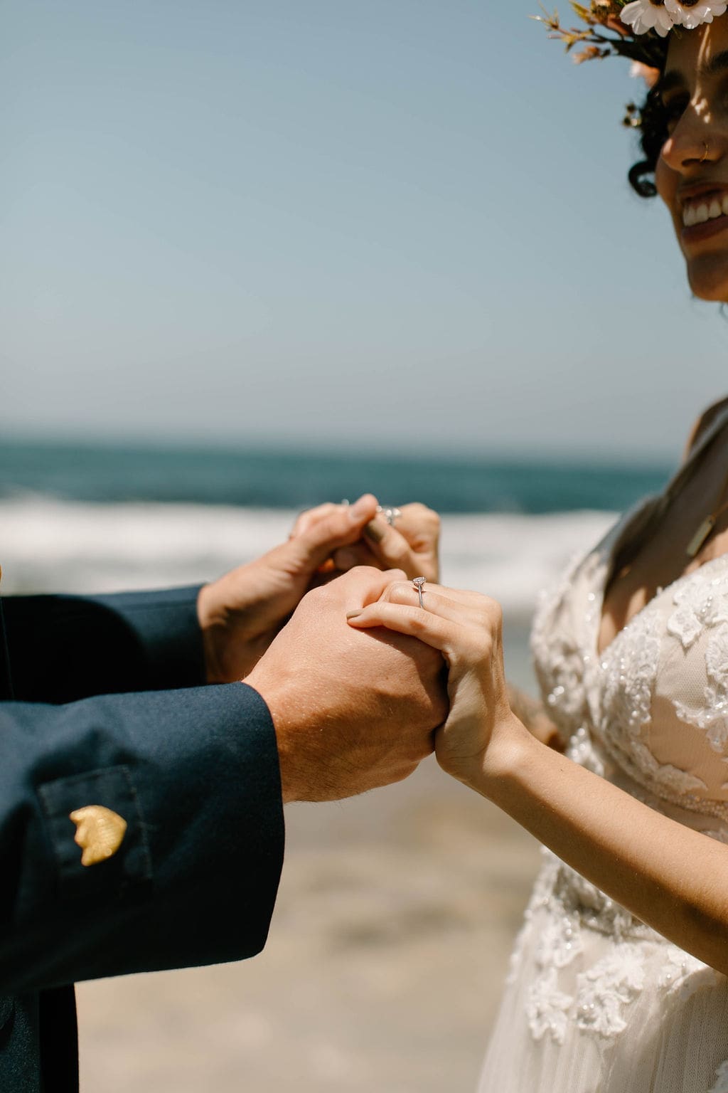 Close-up of hands clasped together, rings glinting in the sun, ocean blurred softly behind — a small, intimate detail from their California elopement.