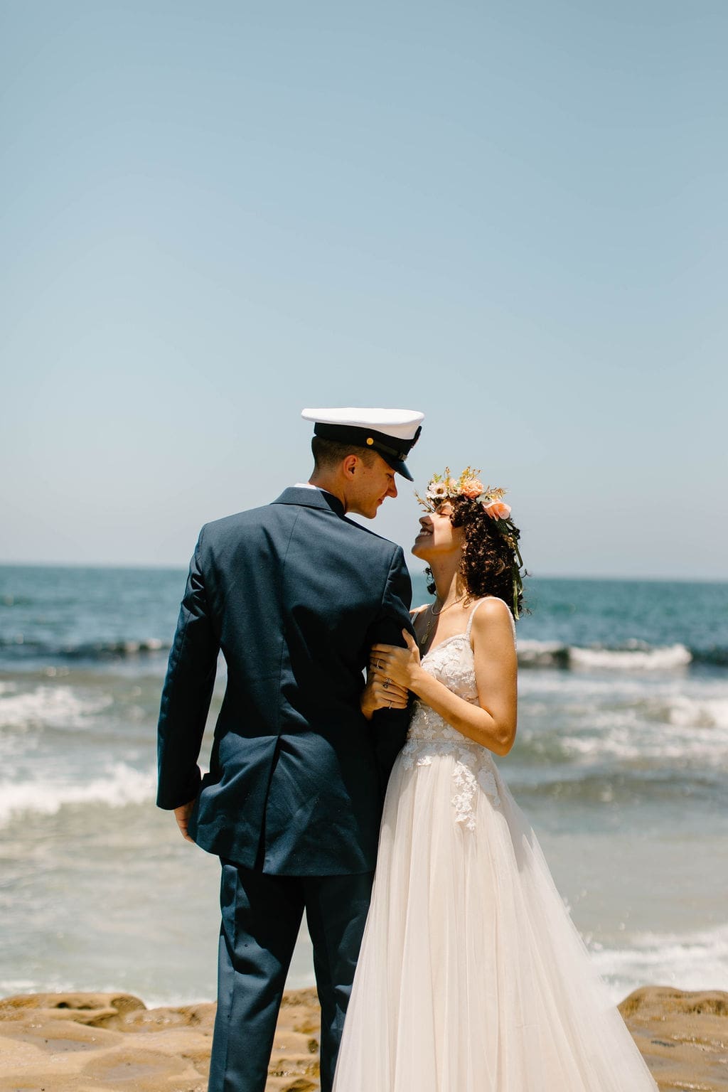 Bride and groom in Navy dress whites lean forehead to forehead laughing on the beach, a candid California elopement moment with the surf rolling in behind them.