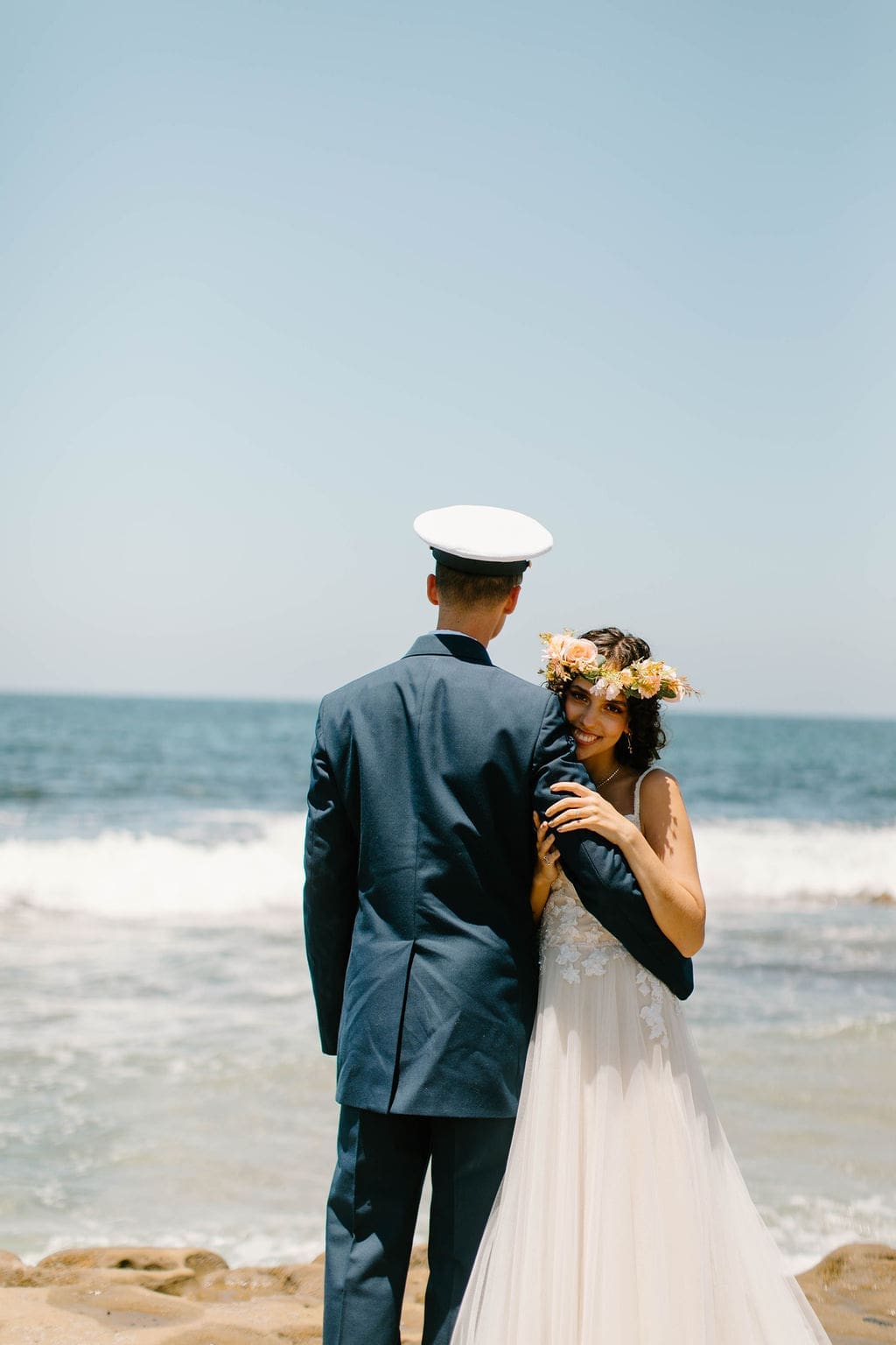 Bride rests her cheek on her groom's shoulder with a bright smile, ocean waves rolling in behind them.