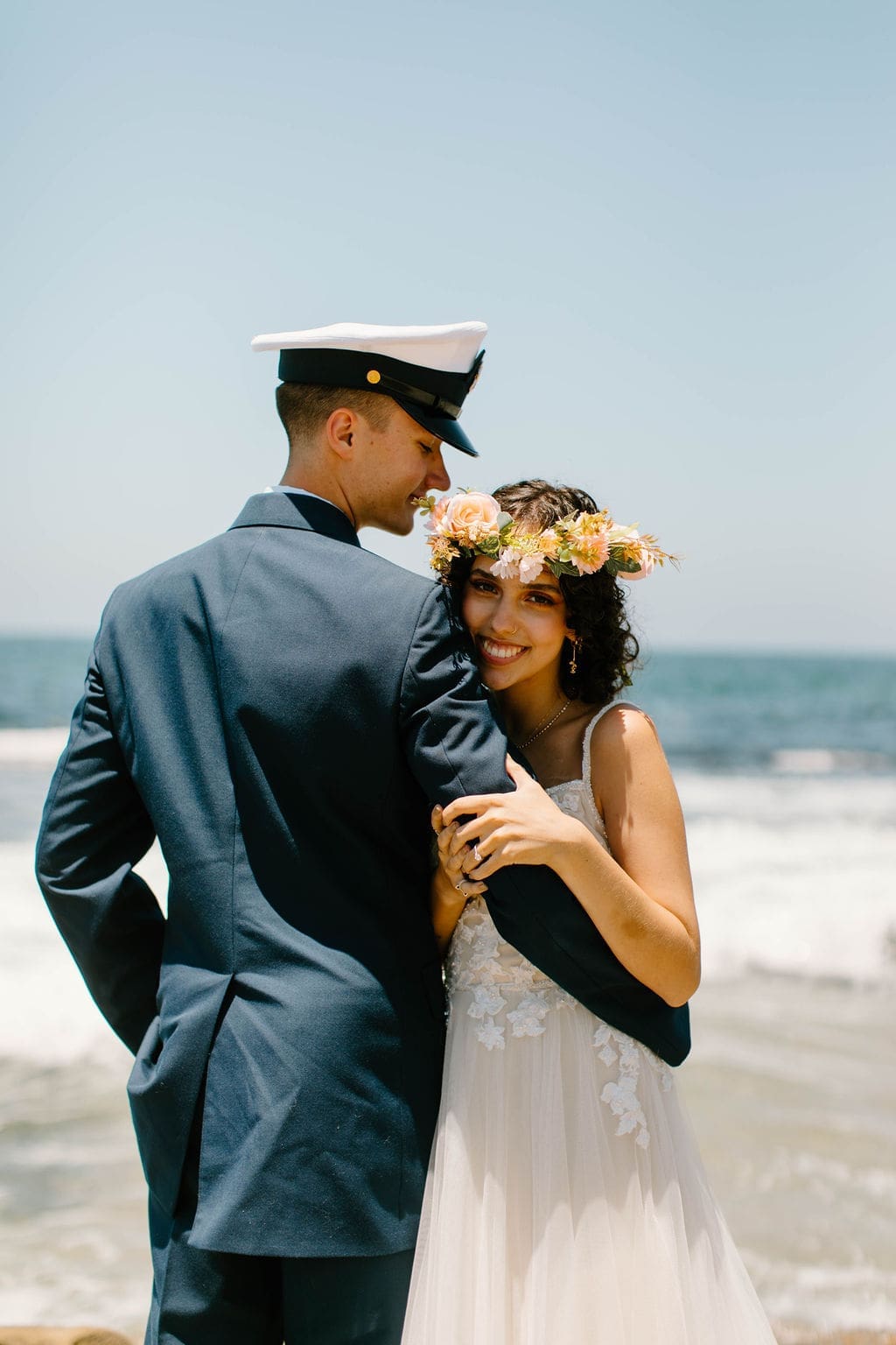 Groom smiles with his eyes closed as his bride nuzzles into his neck, her flower crown catching the light against the sky.