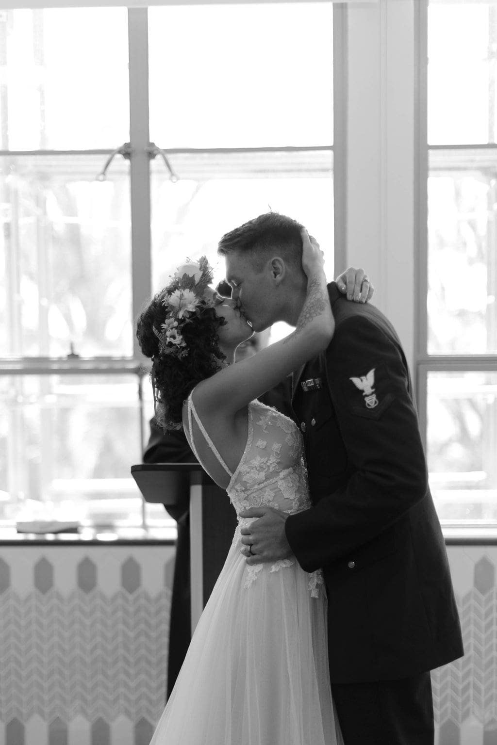 Black and white first kiss at the altar, officiant softly blurred in the background near tall windows.