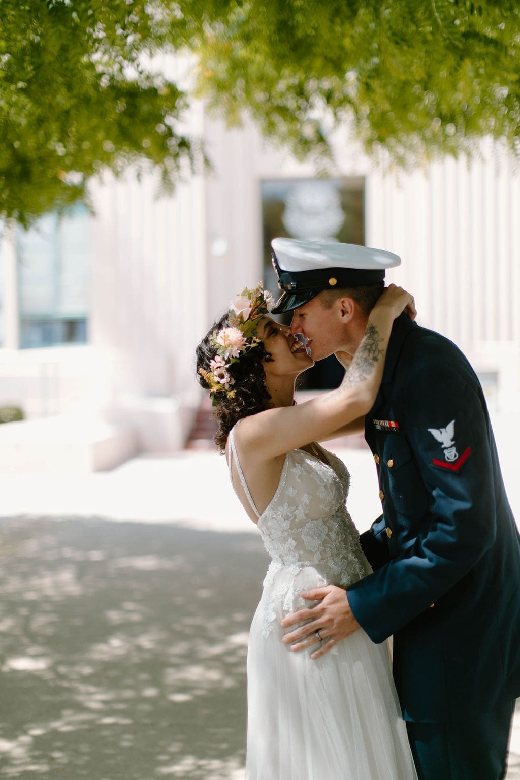Bride and groom share a laughing kiss under dappled tree shade outside a sunlit courthouse-style building, a lighthearted beat in their California elopement.