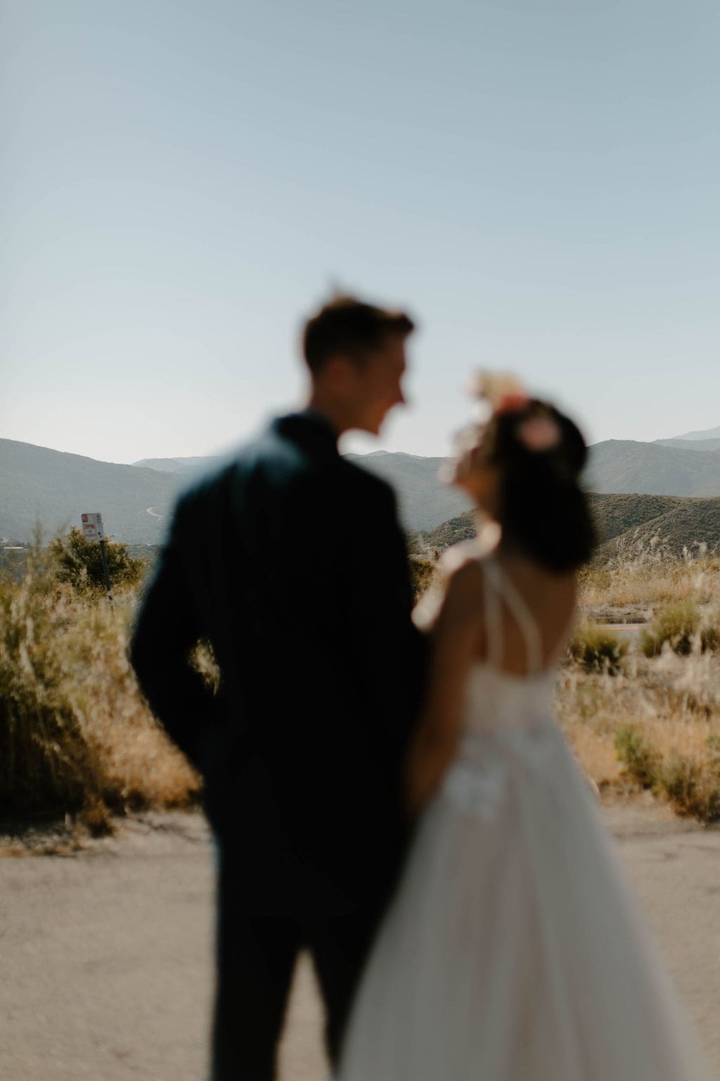 Soft-focus silhouette of bride and groom chatting on a dusty mountain road, hazy hills rolling out behind them.