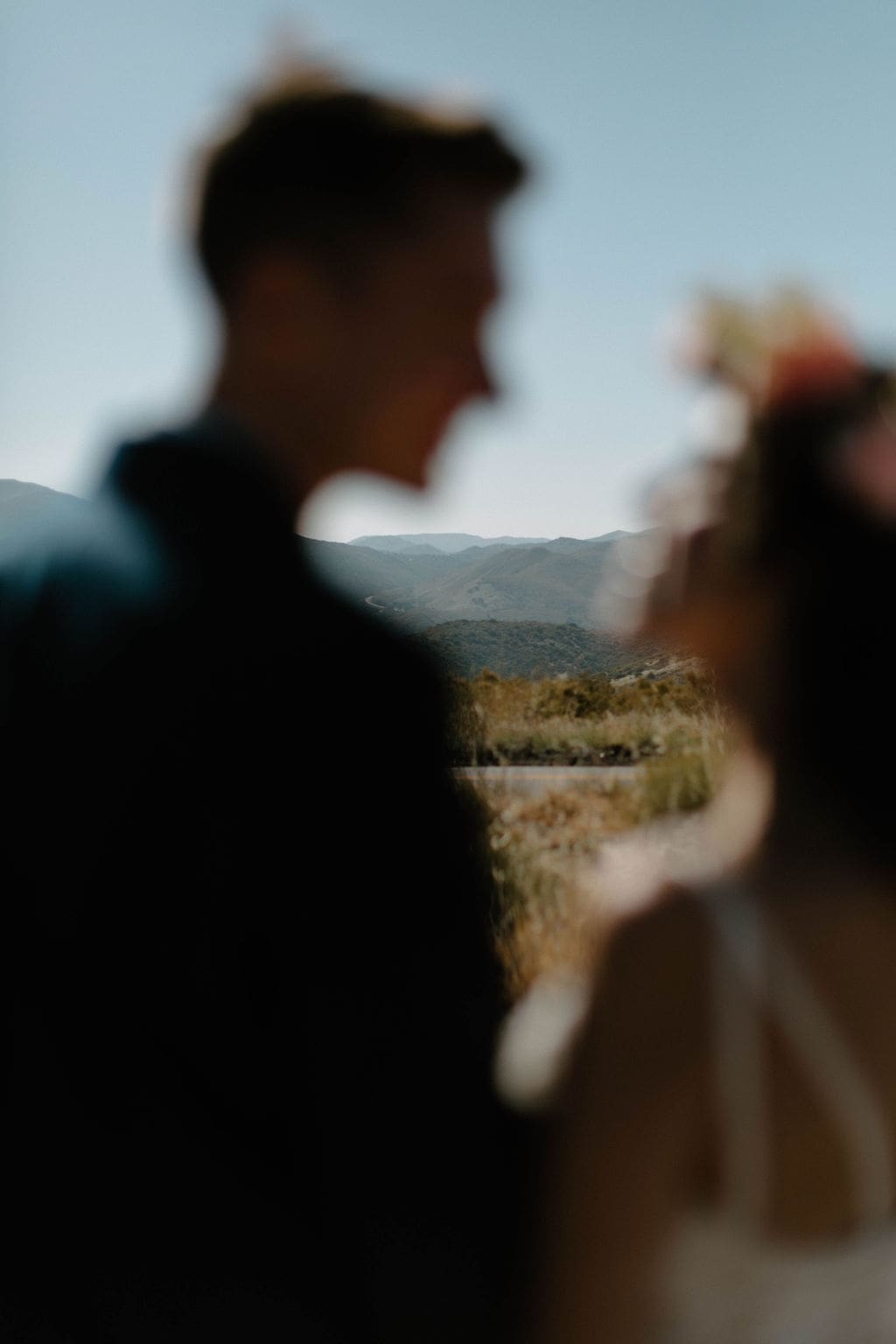 Soft focus shot of the groom laughing with his bride's flower crown blurred in the foreground, mountains layered behind them.