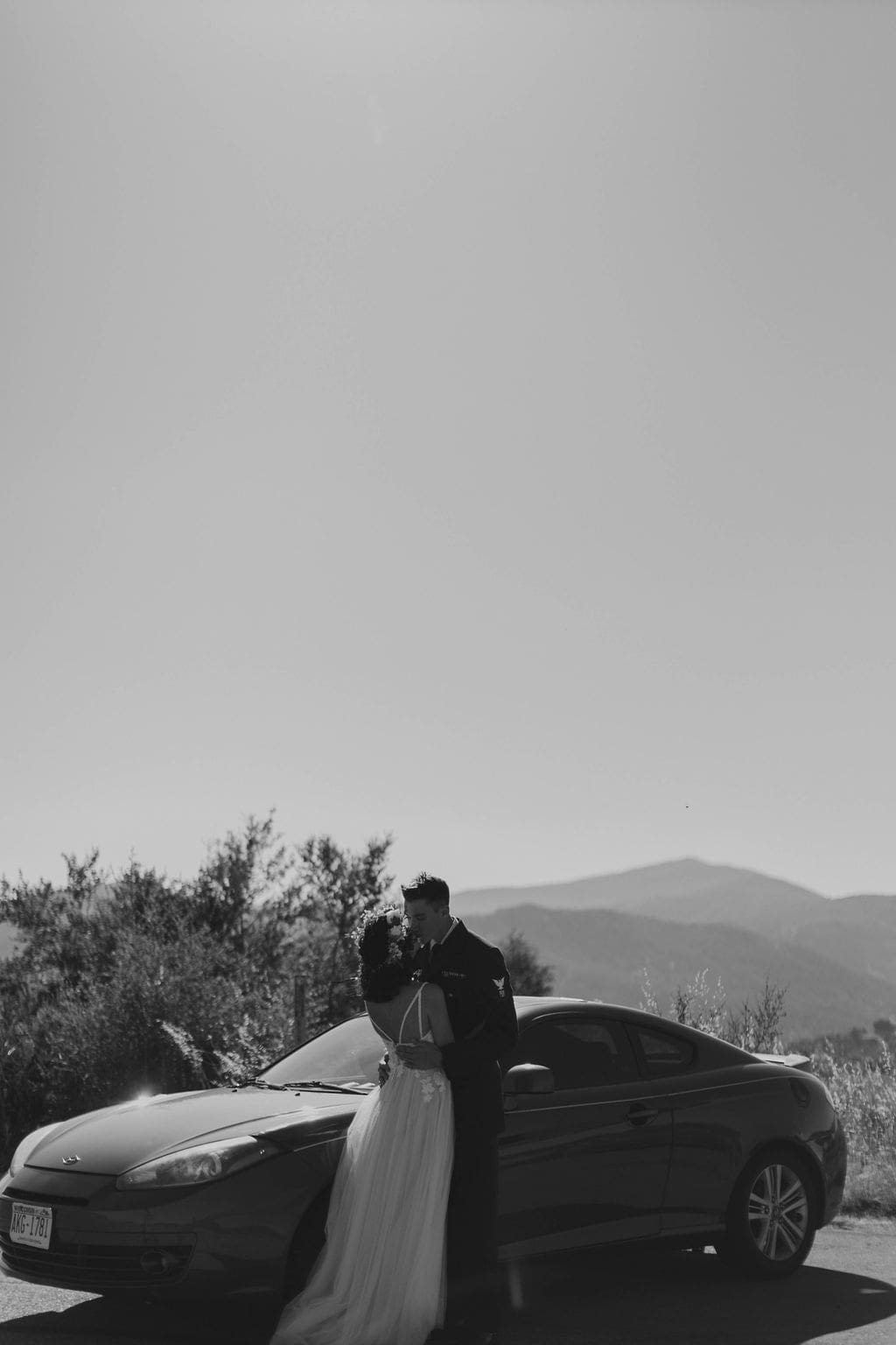 Black and white shot of the couple kissing beside their car, mountain silhouettes fading into the dusk sky.