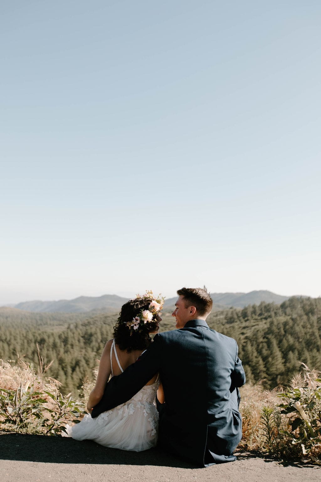 Seated side by side on a ledge, backs to the camera, taking in a sweeping forested mountain view — a peaceful pause during their California elopement.