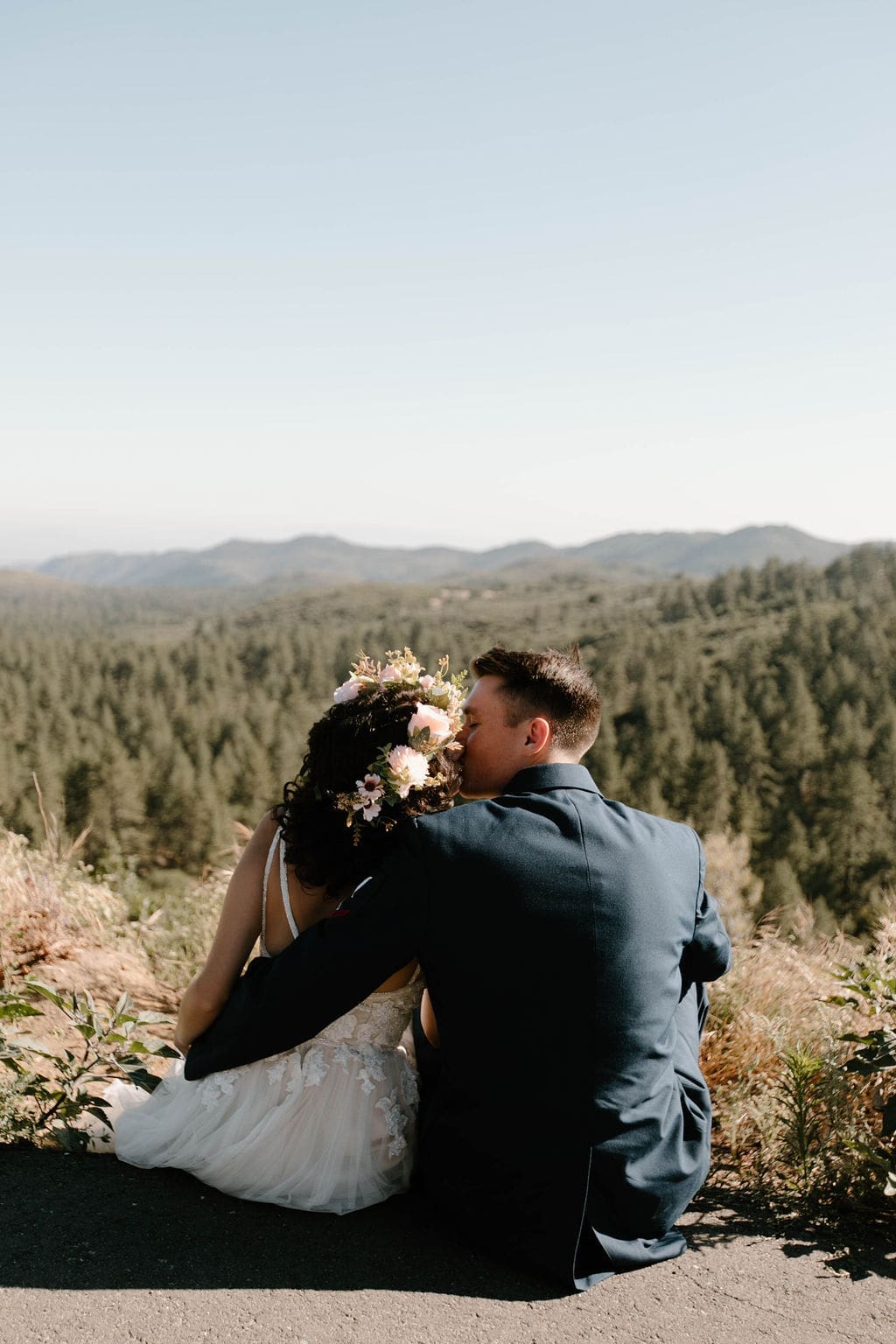 Bride and groom seated together on a ledge, kissing as they look out over a pine covered mountain valley.