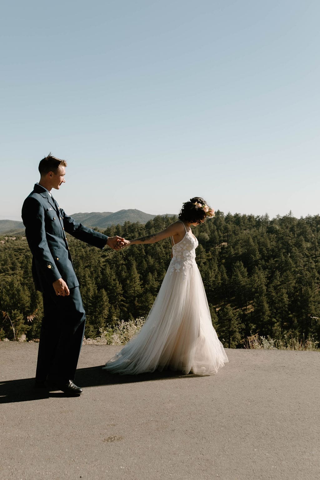 Groom leads his bride by the hand along a scenic overlook, dense forest filling the background.
