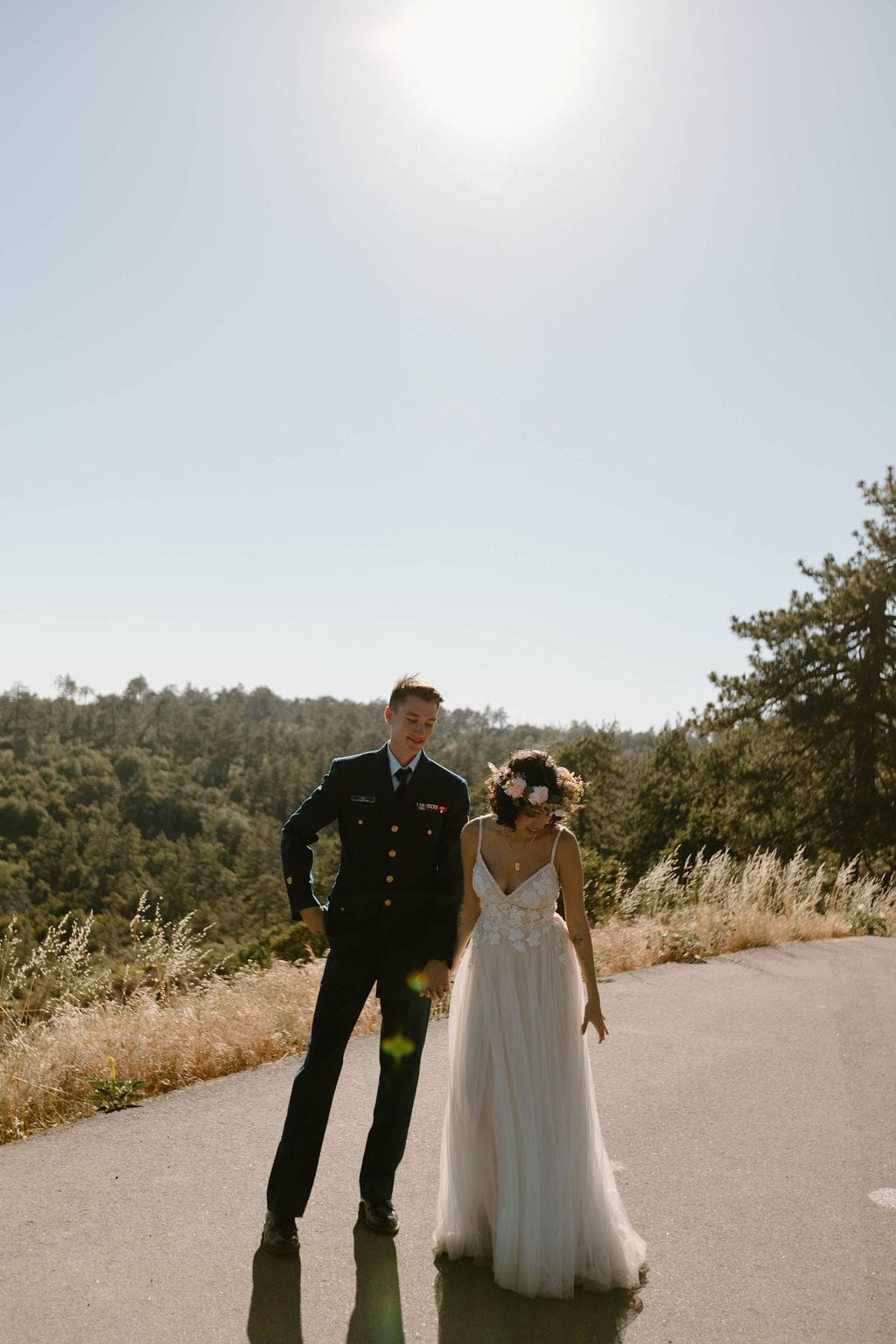 Groom leads his bride by the hand down a sunlit mountain road, her gown trailing behind her.
