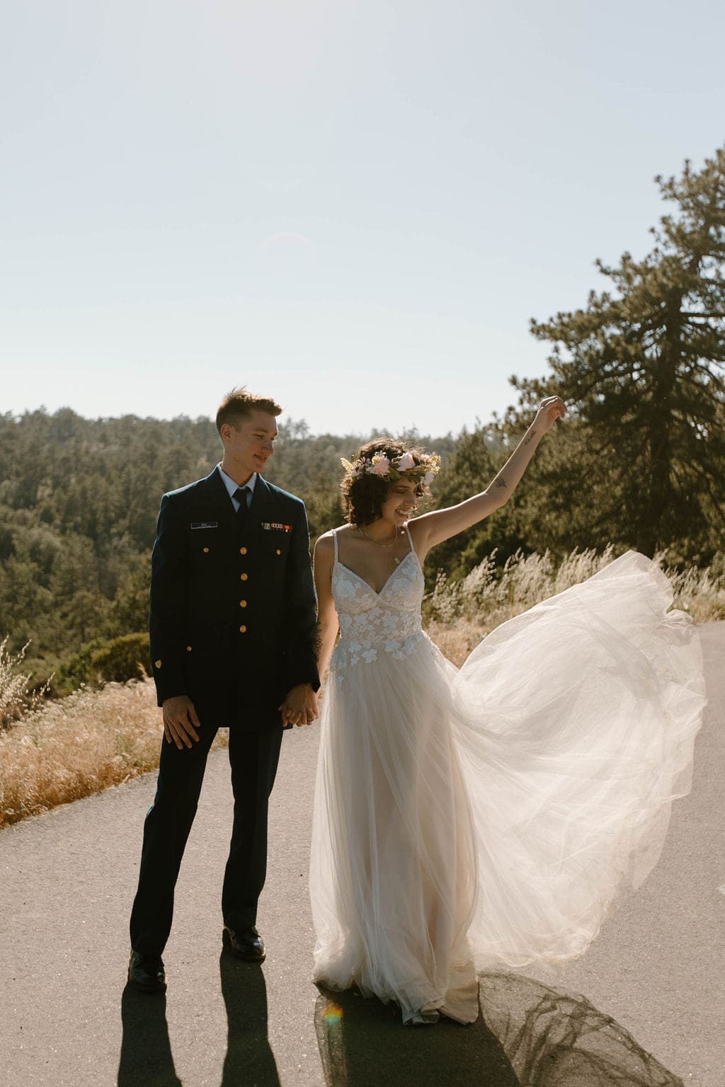 Bride flings her tulle skirt out mid step on the road, laughing beside her groom in his dress uniform, a joyful frame from their California elopement.