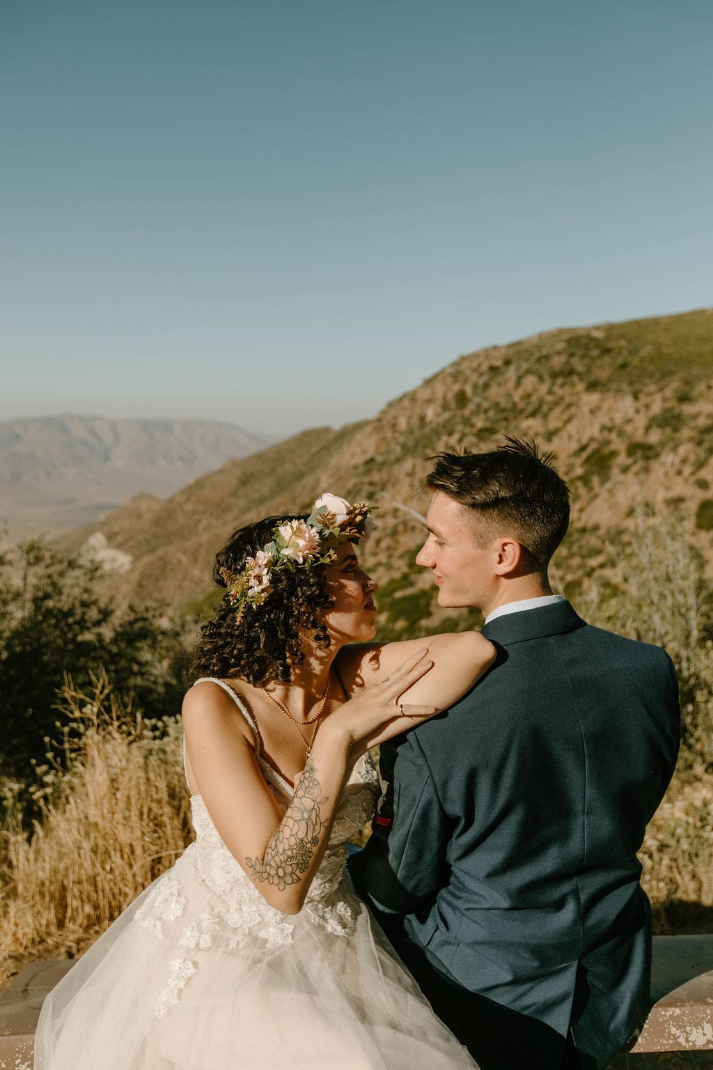 Bride rests her hand on her groom's shoulder while they gaze at each other on a sunlit ridge, dry hills rolling out behind them.