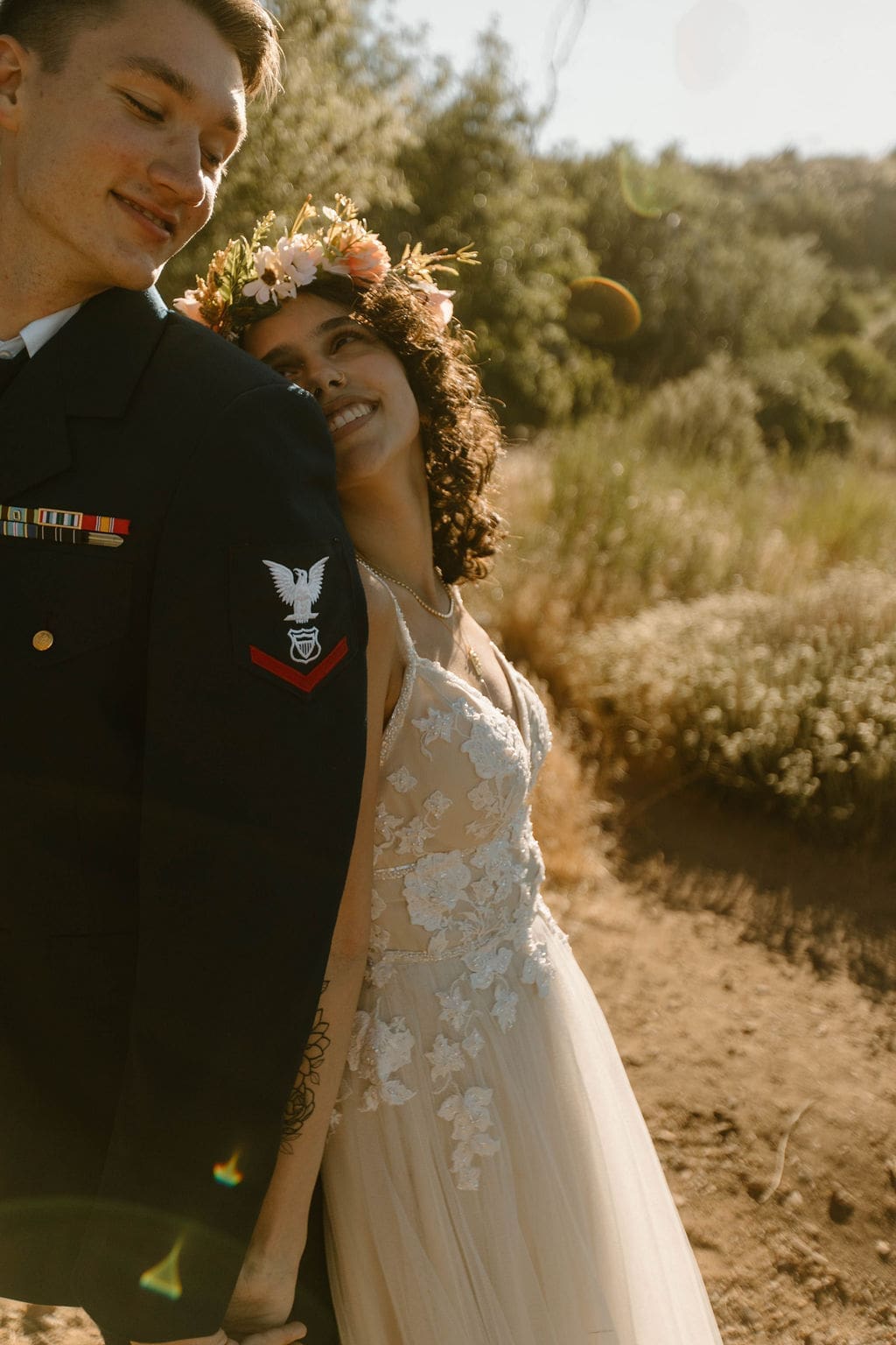 Bride smiles up at her groom's shoulder in golden hour light, his tattoo and service ribbons visible on his sleeve.