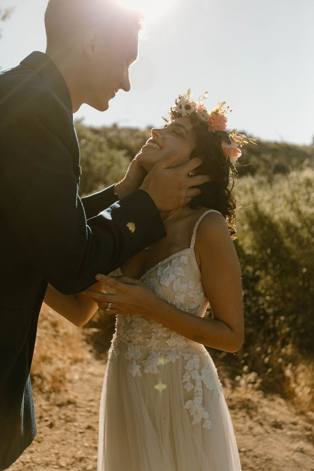 Groom holds his bride's face gently as she laughs, golden hour light washing over the hillside behind them.