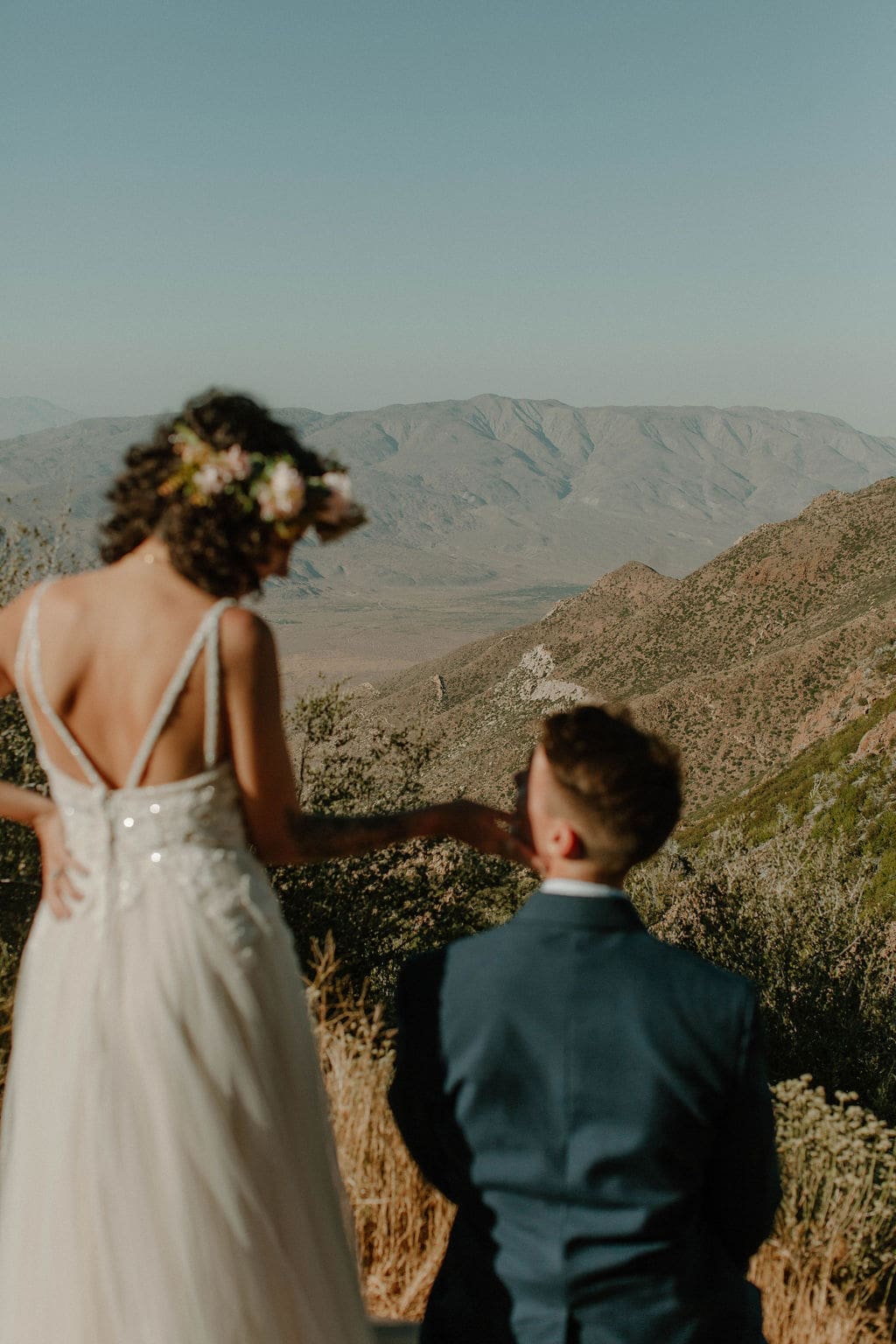 Bride reaches back to touch her groom's chin as they take in a sweeping desert mountain view together.
