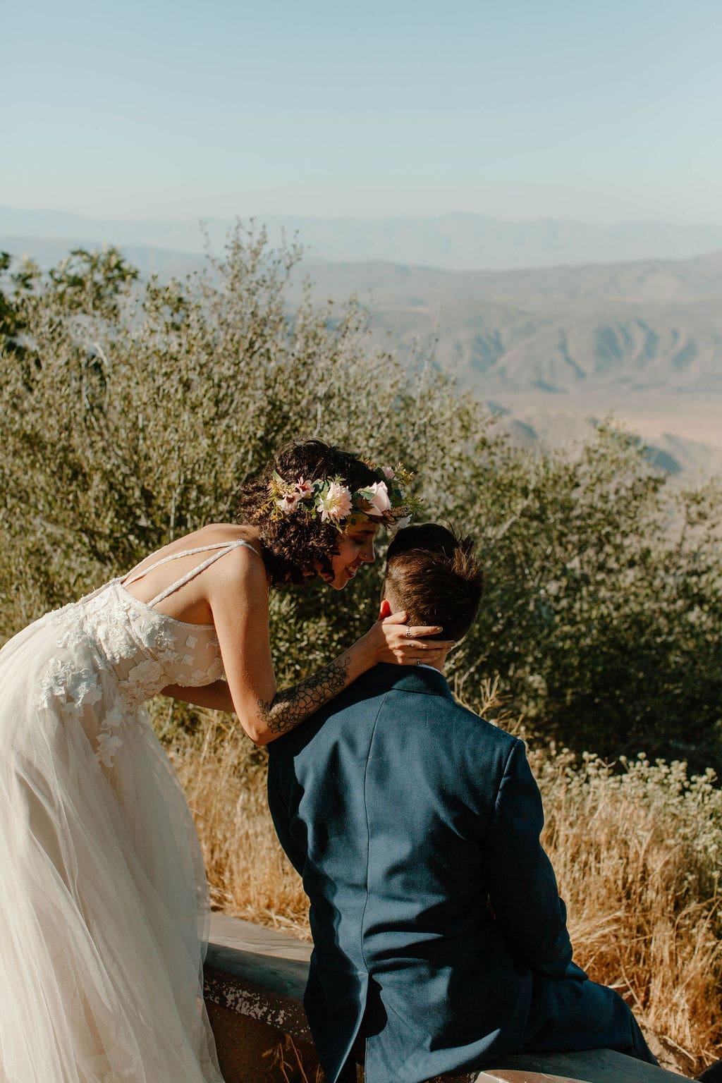 Bride leans down to kiss her seated groom on a bench overlooking a hazy desert valley.