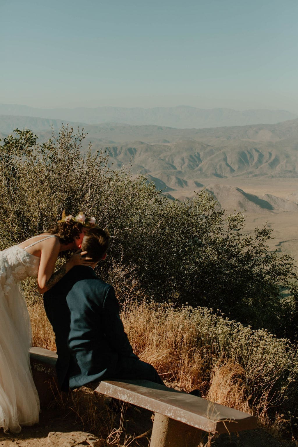 Bride leans down to kiss her groom on a mountainside bench, wildflowers and hazy desert peaks stretching out behind them during their California elopement.