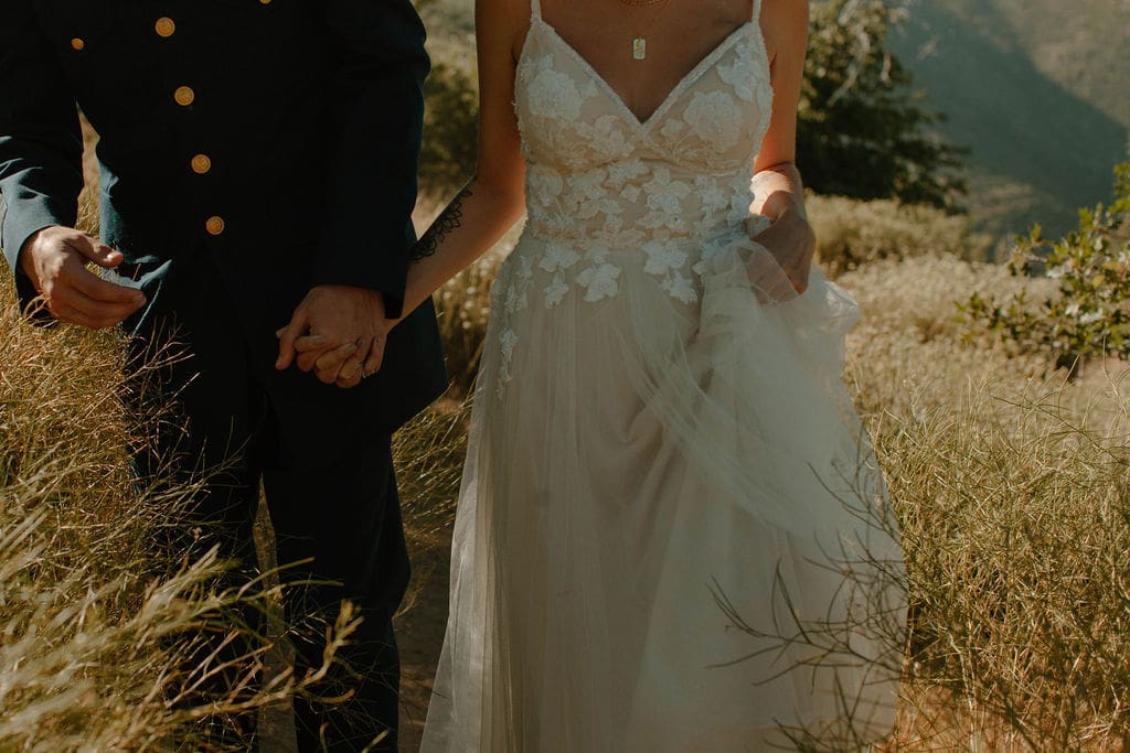 Close crop of the couple holding hands in tall golden grass, her lace bodice and tulle skirt catching the late afternoon light.