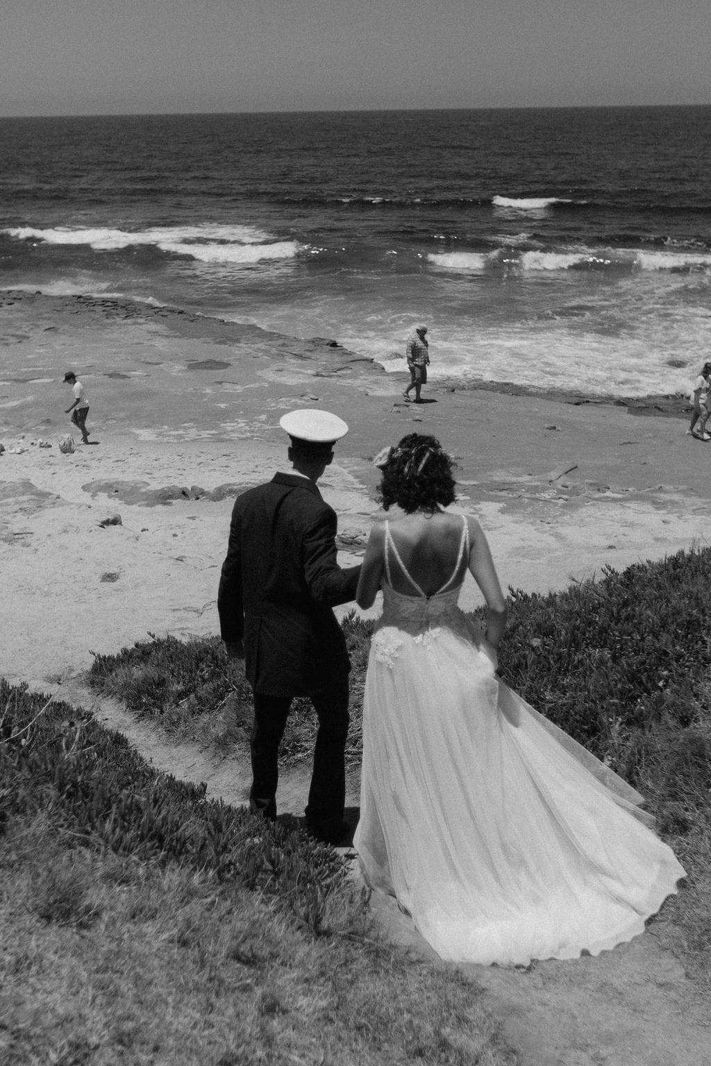 Black and white shot of the couple walking hand in hand down to the shoreline, her dress train trailing behind as beachgoers wander in the distance.