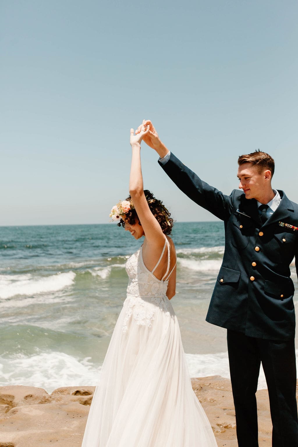 Groom twirls his bride under his arm on the sand, her flower crown and gown catching the sun.