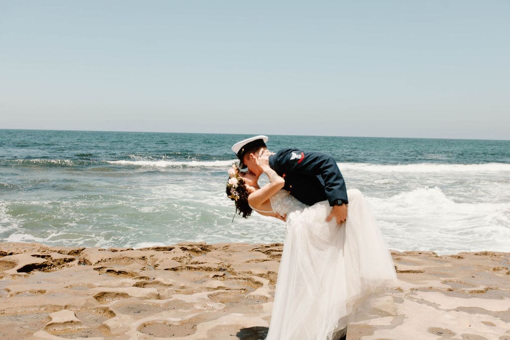 Groom dips his bride for a kiss on the tide-washed rocks, waves crashing just behind them.