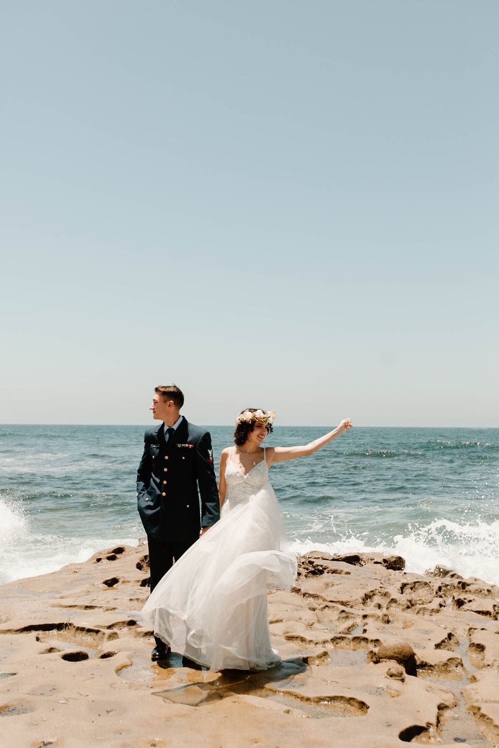 The bride flings her arm out mid-laugh on the rocky shoreline, groom beside her in his dress blues — pure joy captured during their California elopement.