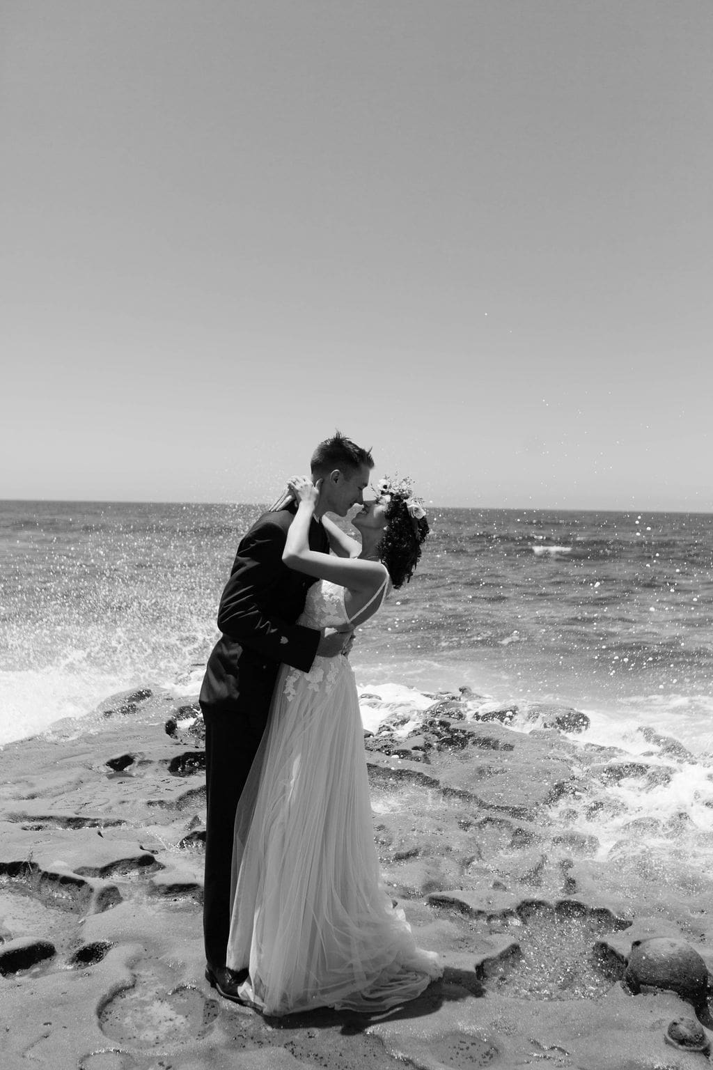Black and white beach kiss, groom holding his bride close as the tide rolls in behind them.