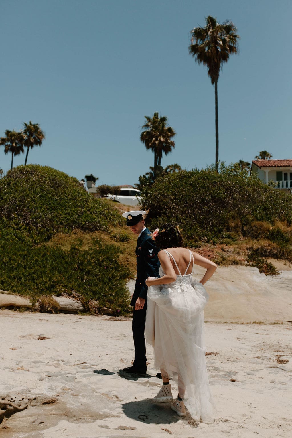 Bride fixes the hem of her gown on the sand with palm trees and a coastal cottage behind her, groom waiting patiently in uniform.