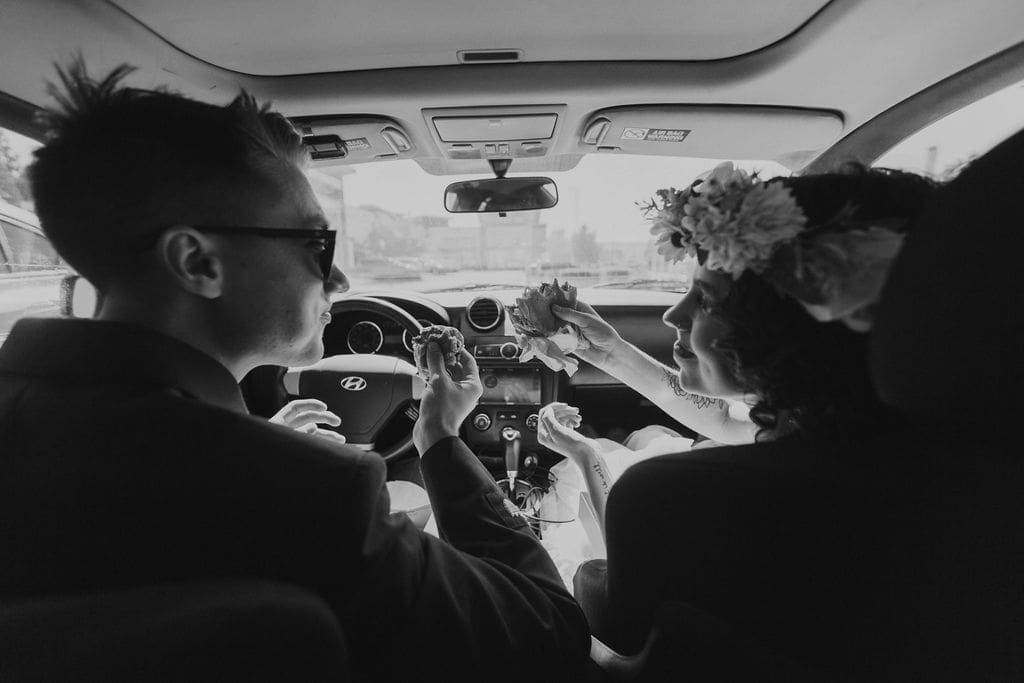 Black and white candid inside the car, bride in her flower crown feeding her new husband a bite of food as he drives.