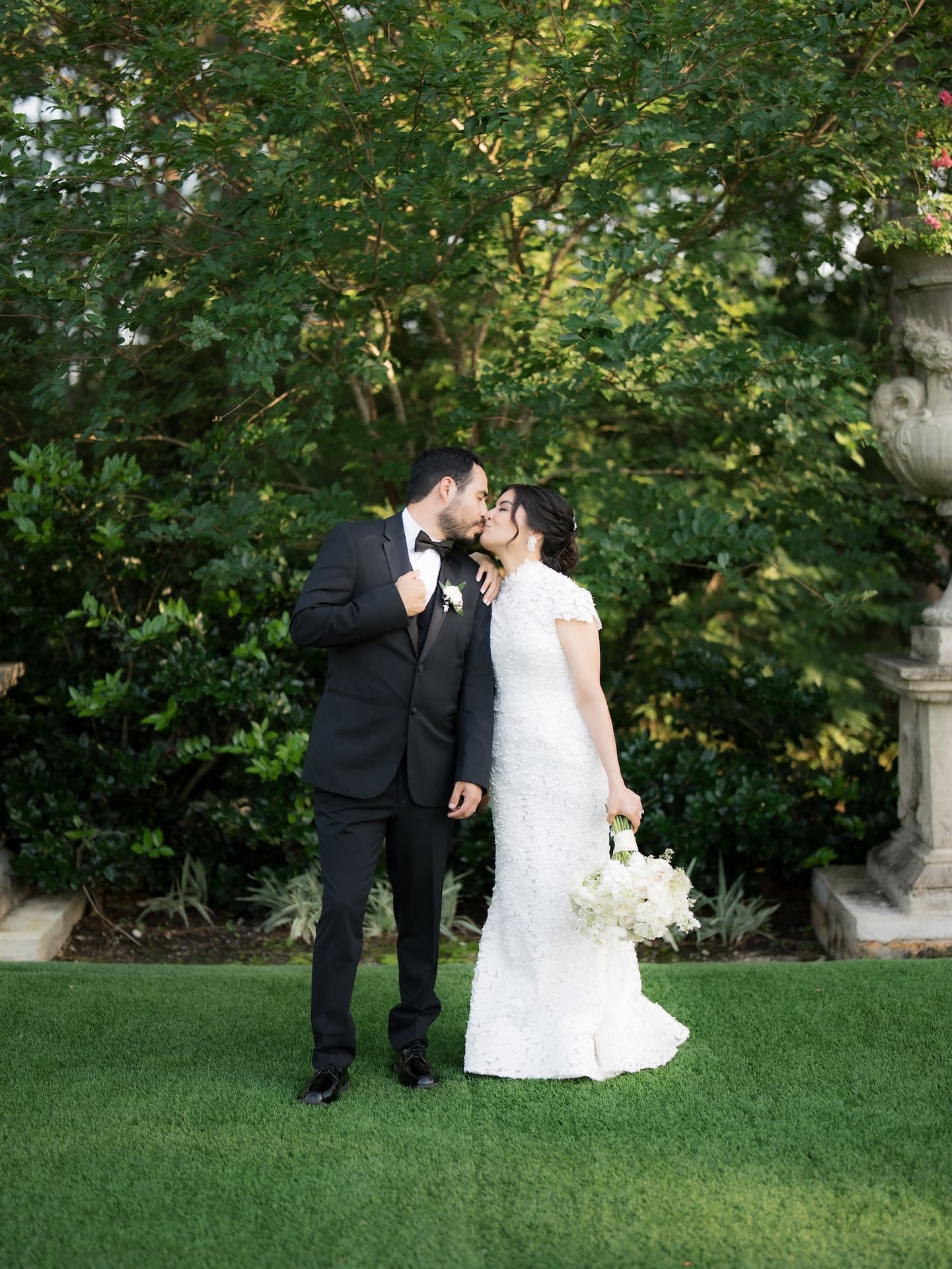 Bride and groom sharing a kiss on manicured green lawn beside a classical stone urn, groom in black tuxedo with white boutonniere.