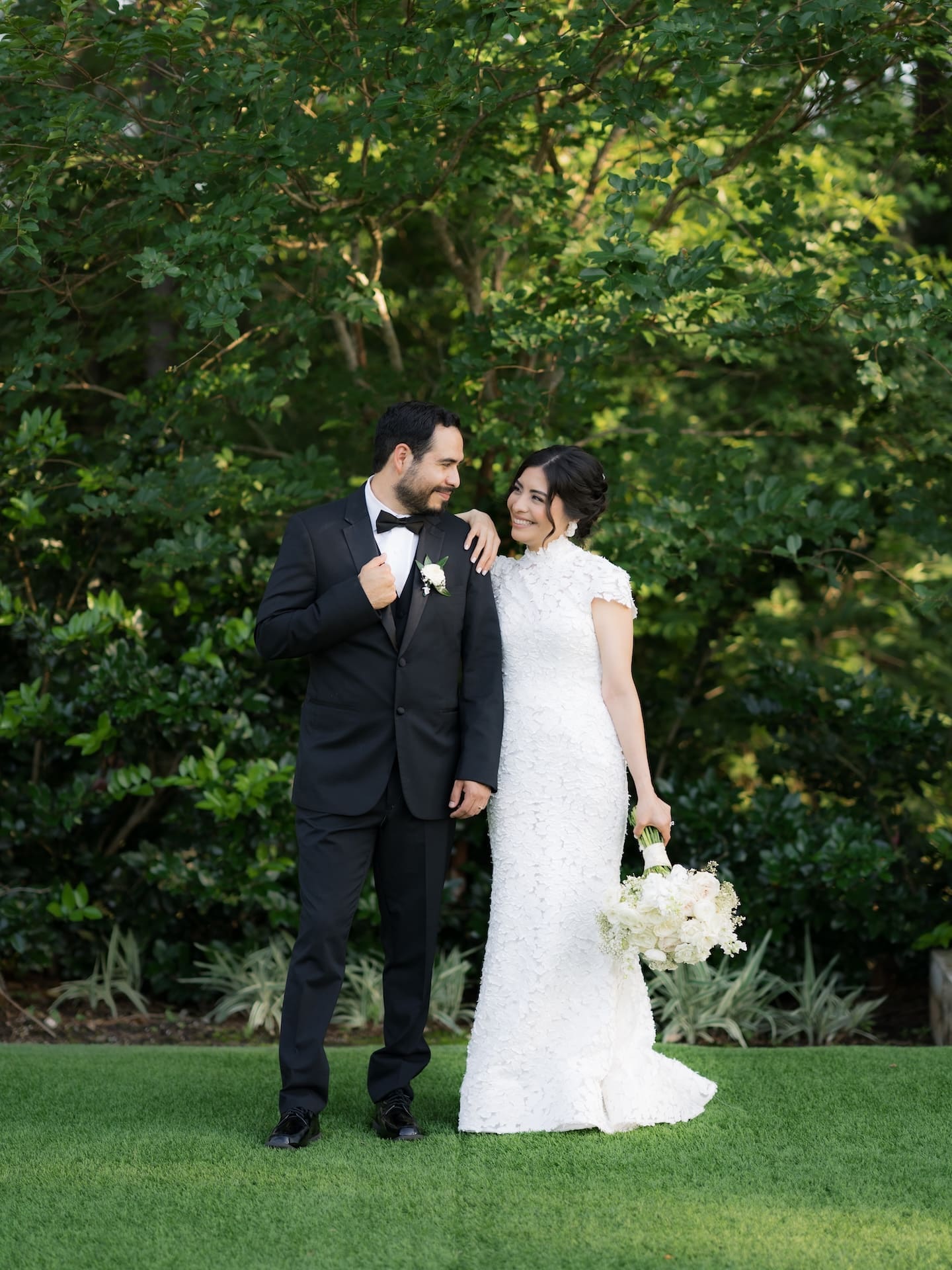 Bride and groom smiling at each other on a green lawn framed by lush trees, groom in a black tuxedo with a white boutonniere.