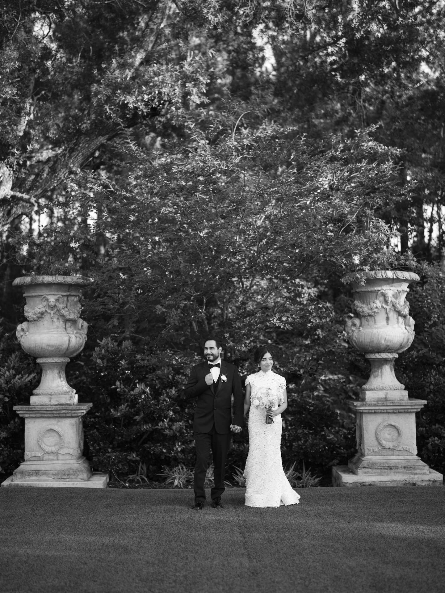 Black and white portrait of bride and groom standing side by side on a lawn between two large stone garden urns and mature trees.