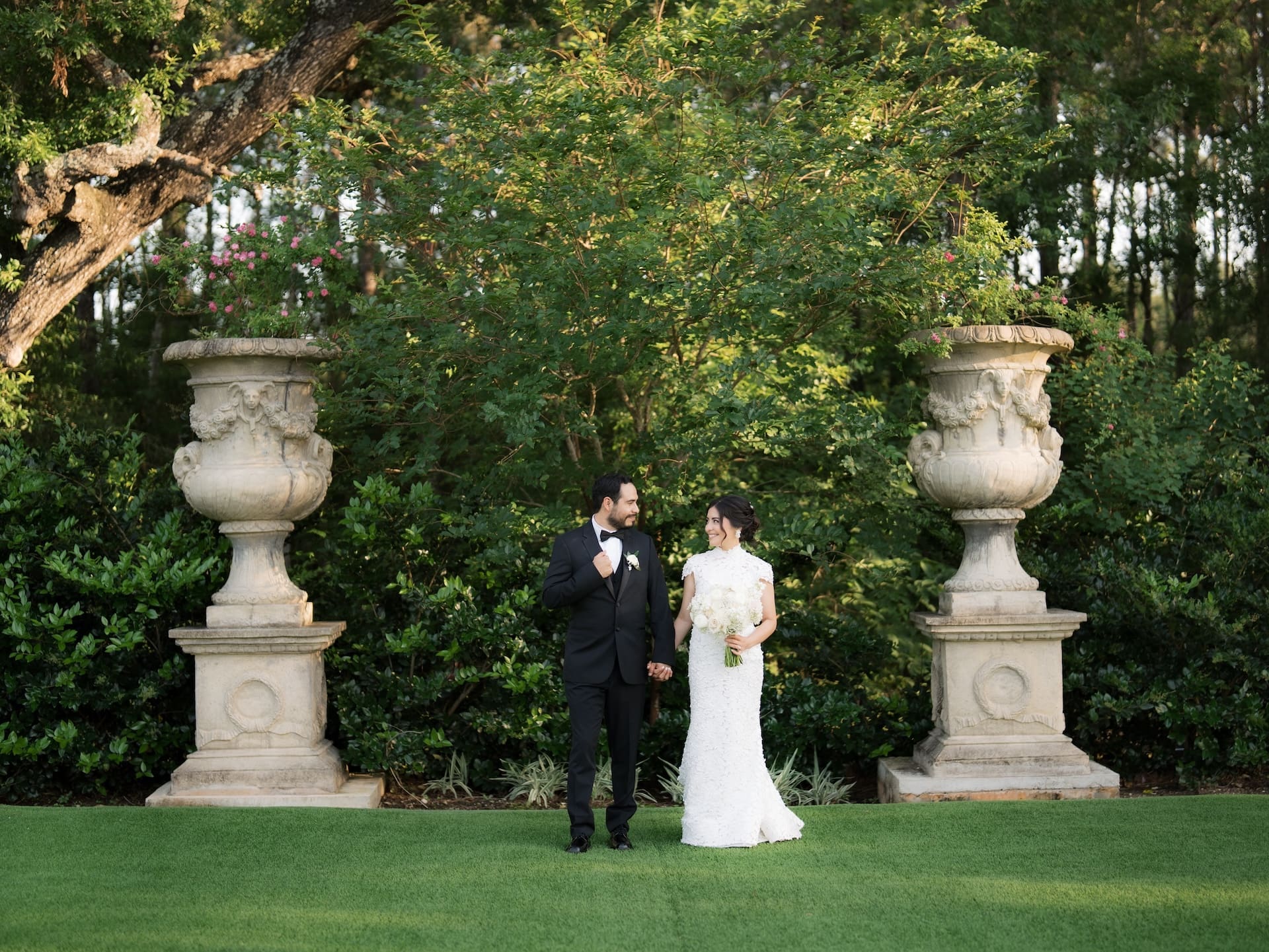 Bride and groom holding hands and smiling at each other on a green lawn, flanked by tall classical stone urns and lush trees.