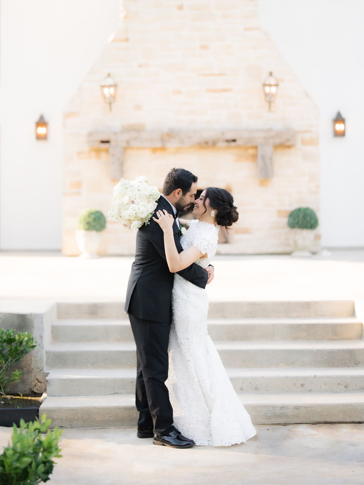 Bride and groom embracing nose-to-nose on outdoor steps in front of a stone fireplace, her bouquet of white roses tucked between them — a tender moment from this luxury wedding.
